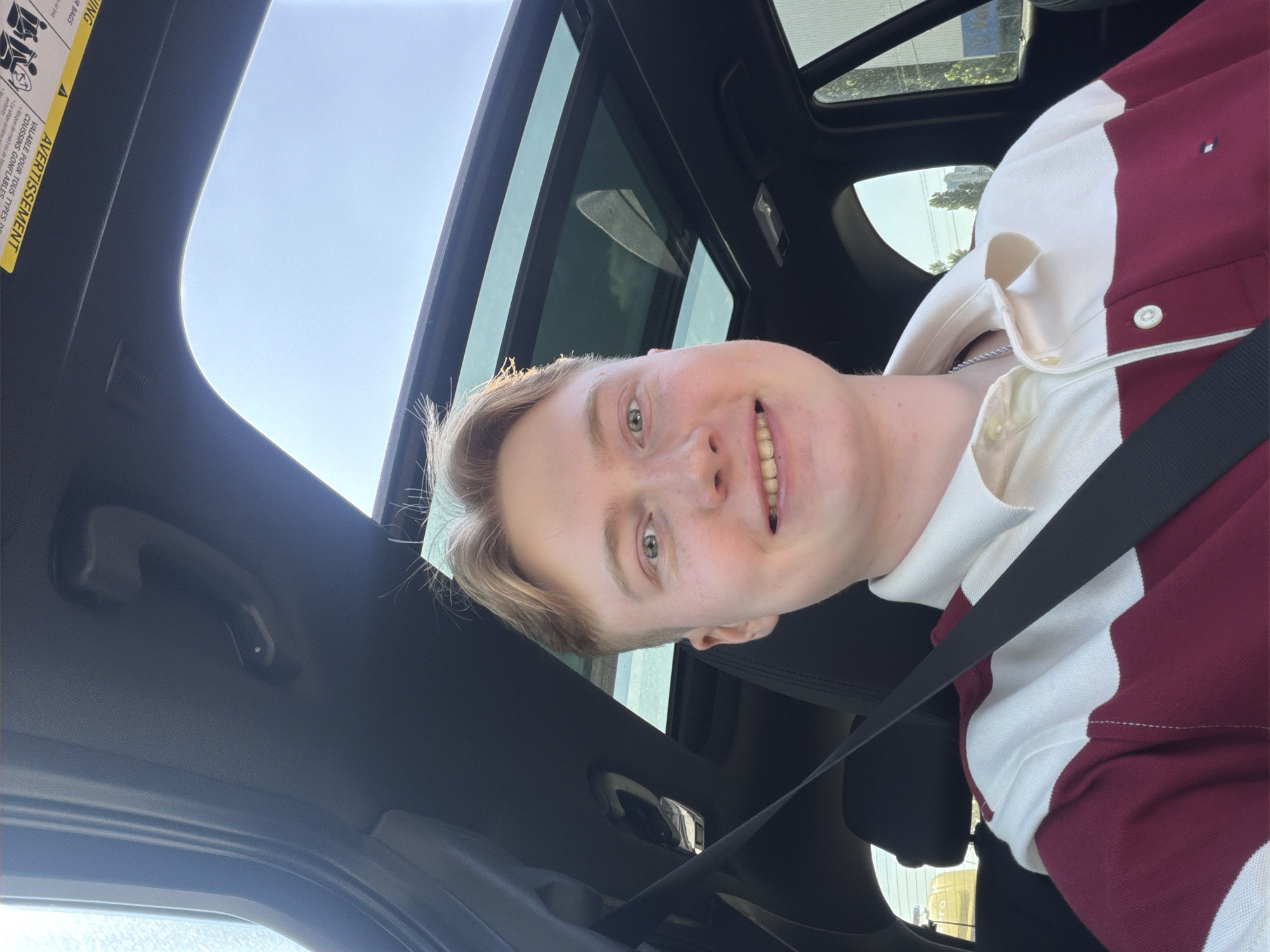 A young Caucasian male with light brown hair smiles at the camera while sitting in a car. He is wearing a long-sleeved collared shirt with maroon and white horizontal stripes. A black seatbelt crosses his chest. The car has a panoramic sunroof, through which the sky and some trees are visible. A warning label with French text is affixed to the interior of the car, near the sunroof. The overall lighting suggests it is daytime. There is no information available to determine the specific location within the city of Kyiv, Ukraine.