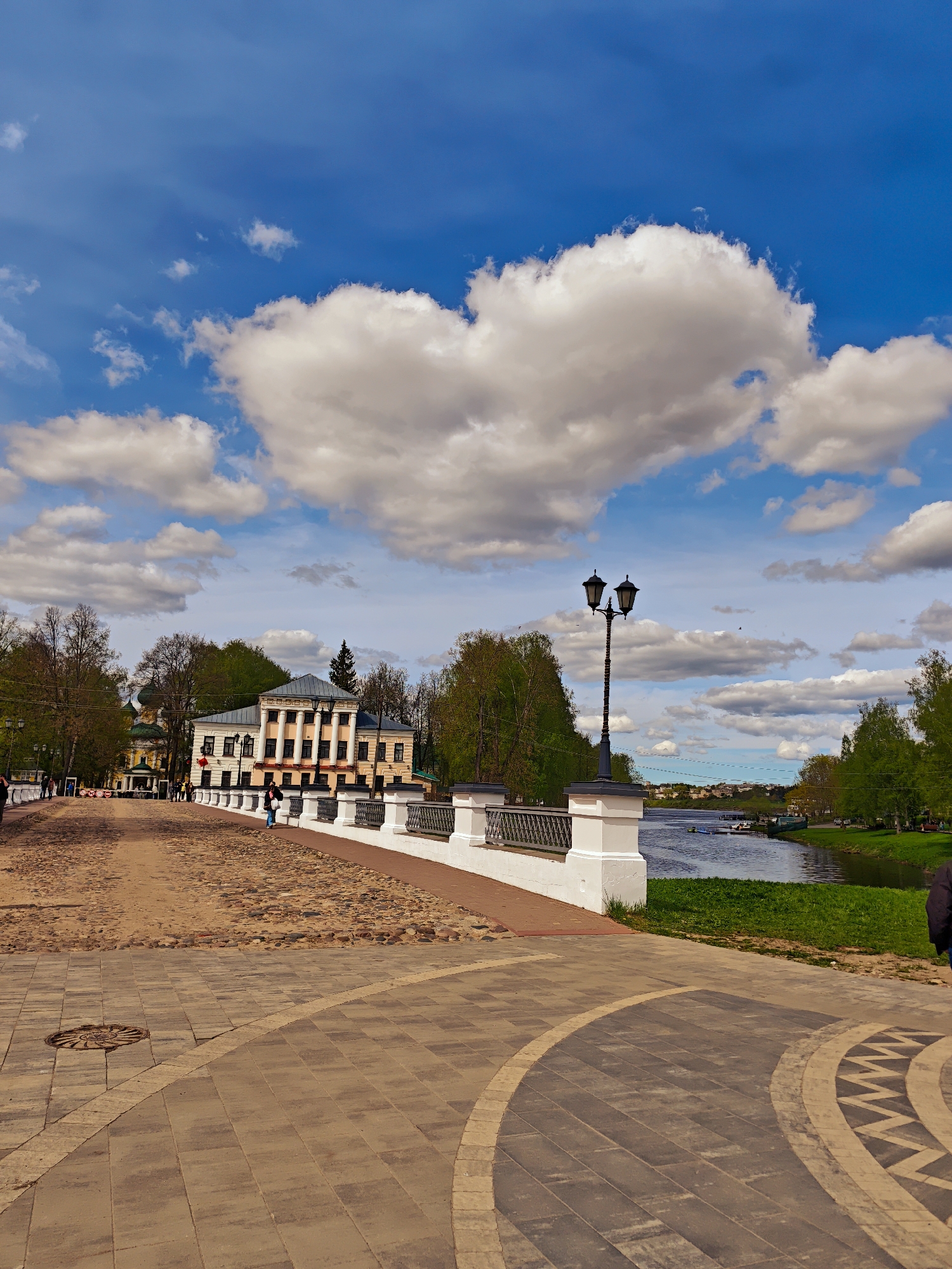 The image shows a view from a cobbled bridge in Uglich, Russia, under a partly cloudy sky. A prominent white cloud in the sky resembles a heart shape. A two-story historical building with a colonnaded facade stands to the left of the bridge. A black lamppost is located on the bridge near the river. The bridge has a white railing. There are people walking on the bridge and along the riverbank. The river flows to the right, with greenery along its banks. The foreground shows patterned paving stones.