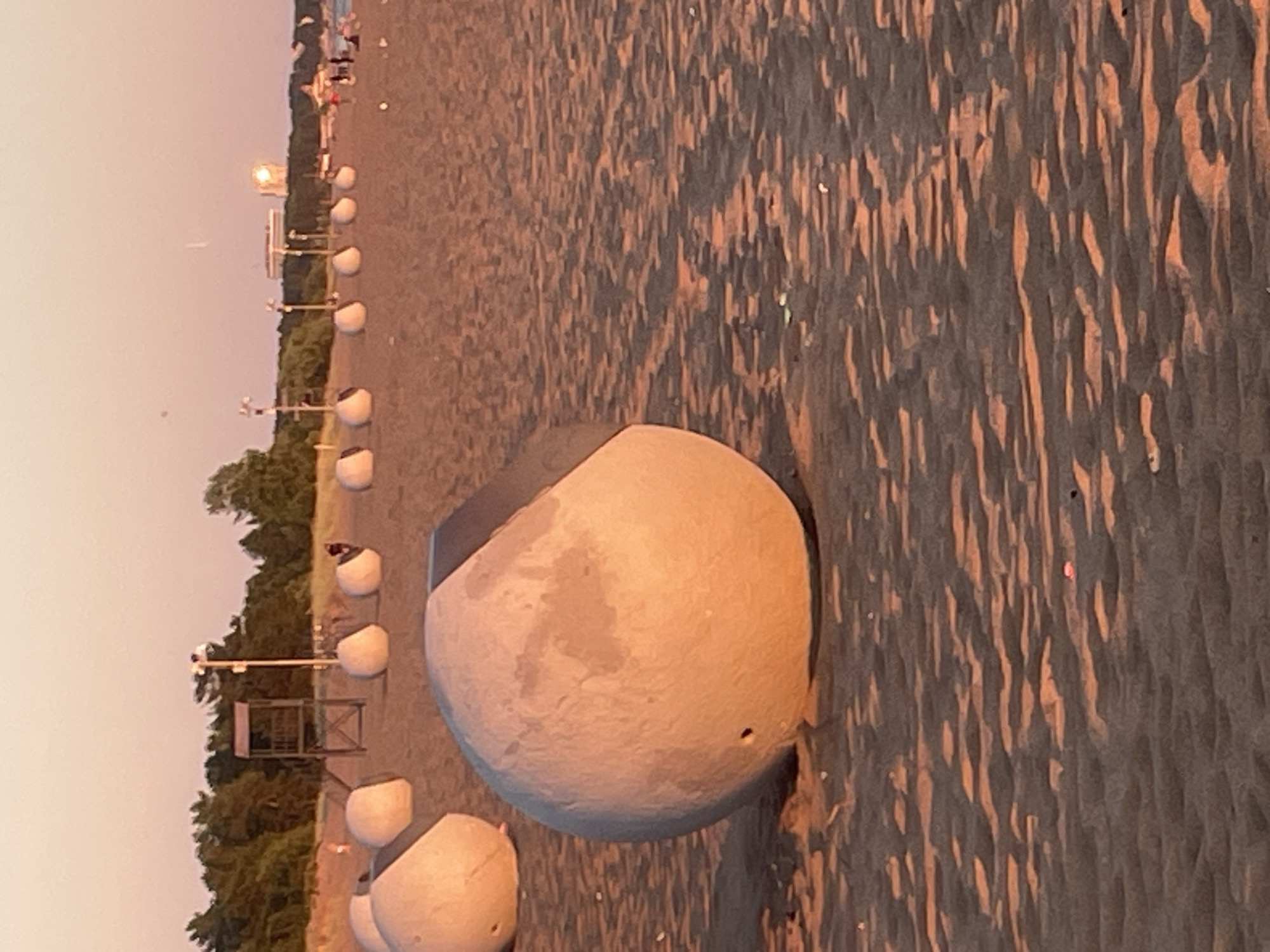 The image shows a beach at sunset in Solnechnoye, Russia. In the foreground, there are large, light-colored, spherical concrete objects on the sand. One of these objects is prominently featured and is partially cut off at the top. In the middle ground, more of these spherical objects are arranged in a line. Beyond the spheres, a lifeguard tower stands on the sand, backed by a line of trees. In the distance, buildings and more trees are visible, with a bright glare in the sky suggesting the setting sun. Several people are scattered on the beach and near the water in the background. The sand is textured with shadows and ripples.