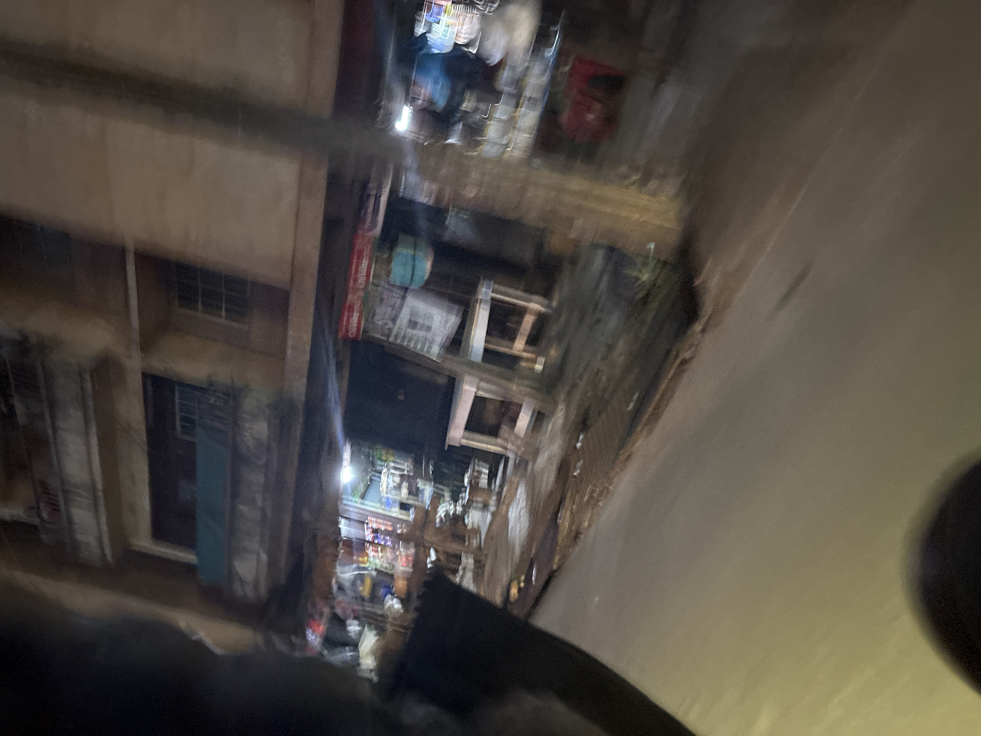 The image depicts a blurred street scene at night in Onitsha, Nigeria. A multi-story building with balconies is visible on the right, showing windows and what appears to be laundry hanging. Below the building, a street-level shop or market area is partially obscured by darkness and motion blur. Various shelves, displays of goods, and what seem to be tables and chairs are present in the shop. A person in a blue top is visible inside the shop, behind a counter. The street in front of the shop is paved and shows a slight incline. A dark, blurred object occupies the left side of the frame, possibly a vehicle or a person. The overall impression is one of a busy, albeit dimly lit, urban commercial area at night.