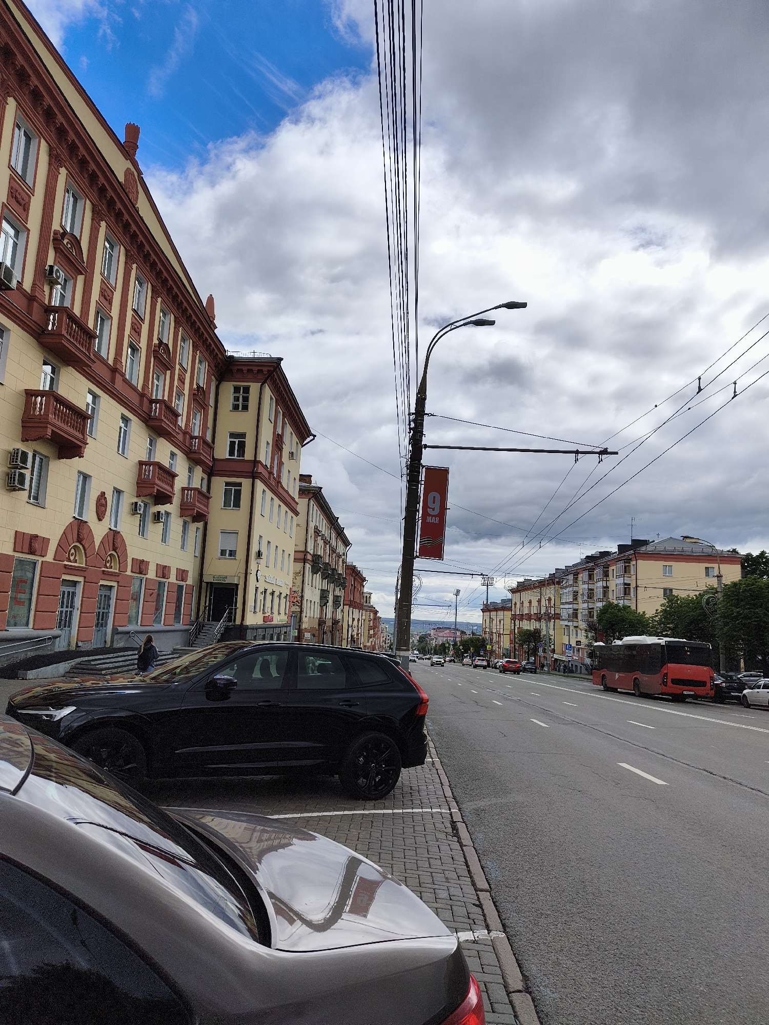 The photograph captures a street scene in Izhevsk, Russia, on an overcast day. The foreground is dominated by the partially visible rear of a gray car and a fully visible black Volvo SUV parked on a cobblestone sidewalk. Behind the vehicles, a wide street stretches into the distance, featuring a red bus on the right side of the road and various other cars scattered along its length. The road is marked with white lines.
On either side of the street are buildings with yellow facades and red-brown trim. The building on the left has architectural details such as balconies and decorative moldings around the windows. A person can be seen entering one of the buildings. Power lines crisscross overhead. A banner attached to a lamppost displays the number "9", presumably related to a date, and "MAR", possibly indicating March. The sky is filled with gray clouds, although patches of blue sky are visible above the buildings on the left. In the distance, faint outlines of hills or elevated terrain are seen.