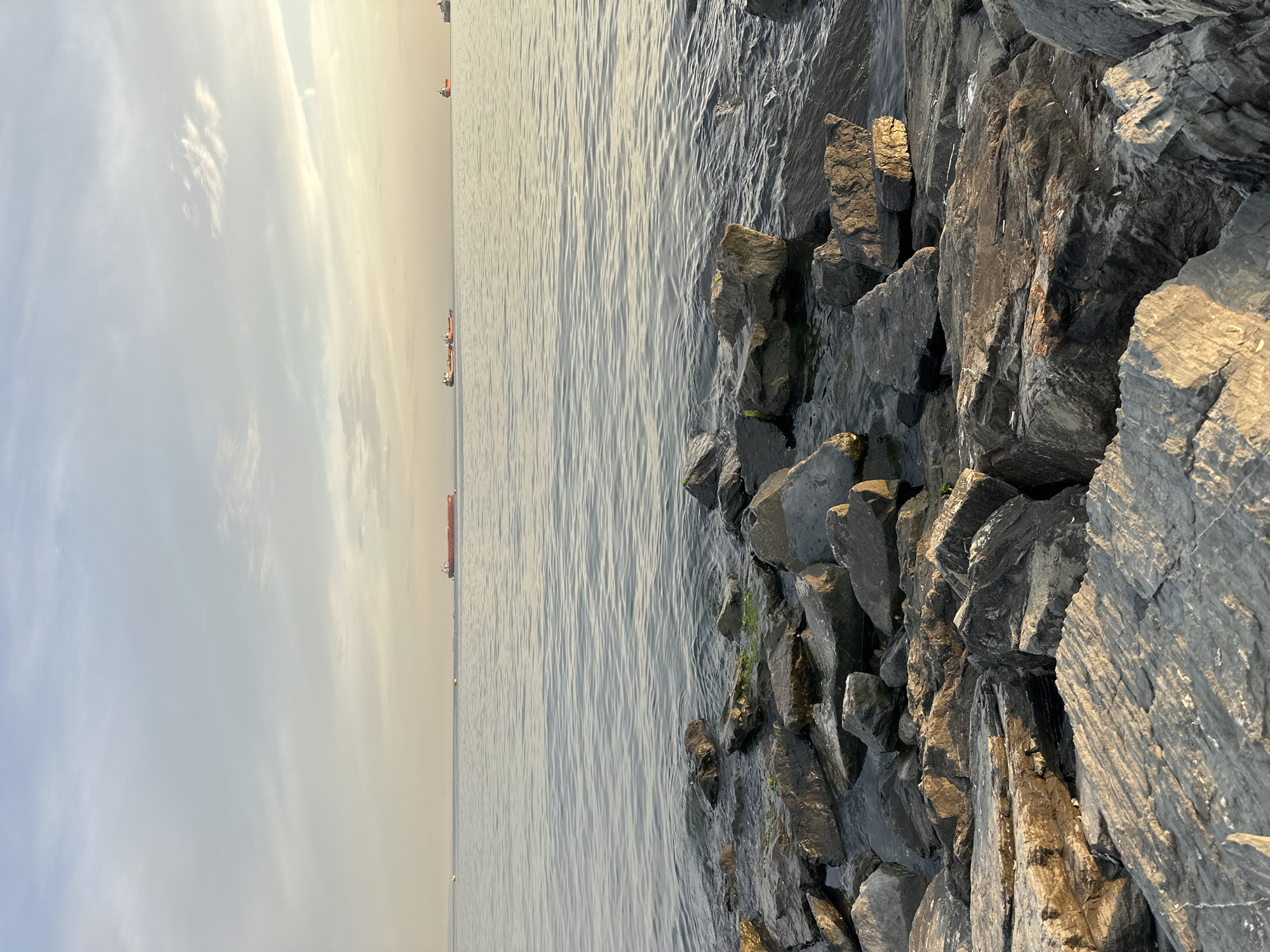 The image shows a rocky shoreline with the sea stretching out to the horizon. Several cargo ships are visible in the distance on the water. The sky is partly cloudy with soft, diffused sunlight, suggesting it is either early morning or late afternoon. The foreground is dominated by large, grey rocks, some of which are wet from the sea. The water is calm with gentle ripples. The overall scene depicts a peaceful coastal environment in Bakırköy, Turkey, likely along the Marmara Sea.