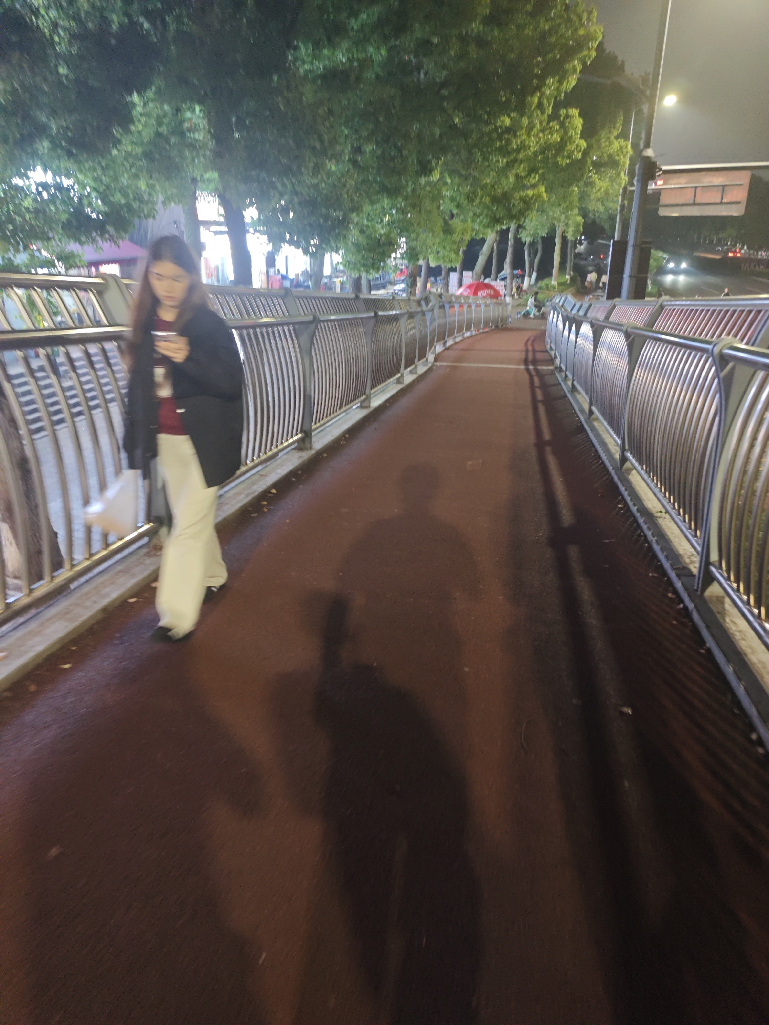 The image depicts a nighttime scene on an elevated pedestrian pathway in Chongqing High-tech Zone, China. A woman is walking on a reddish-brown path, looking down at a smartphone held in her right hand. She is dressed in a dark jacket over a red top and light-colored, possibly white or cream, wide-leg pants, with a light-colored bag in her left hand. Her long, dark hair falls over her shoulders.
The pathway is bordered on both sides by modern, curved metal railings, which are partially reflective. To the left, a line of trees with green foliage is visible, illuminated by artificial light sources from above, creating bright spots. Beyond the trees, in the distance, are faint indications of structures, including a red umbrella and what might be other people or stalls.
Strong, elongated shadows of the photographer and the railings are cast onto the damp-looking path, stretching towards the left and into the distance, suggesting multiple light sources. To the right of the pedestrian path, an adjacent road can be seen, with the blurred headlights of distant vehicles indicating ongoing traffic. A tall pole supporting a dark, rectangular sign or traffic signal is present on the far right, but any text on it is unreadable due to distance and blur. The sky is dark, confirming the nighttime setting. The artificial lighting creates an urban, calm atmosphere, highlighting the modern infrastructure.
