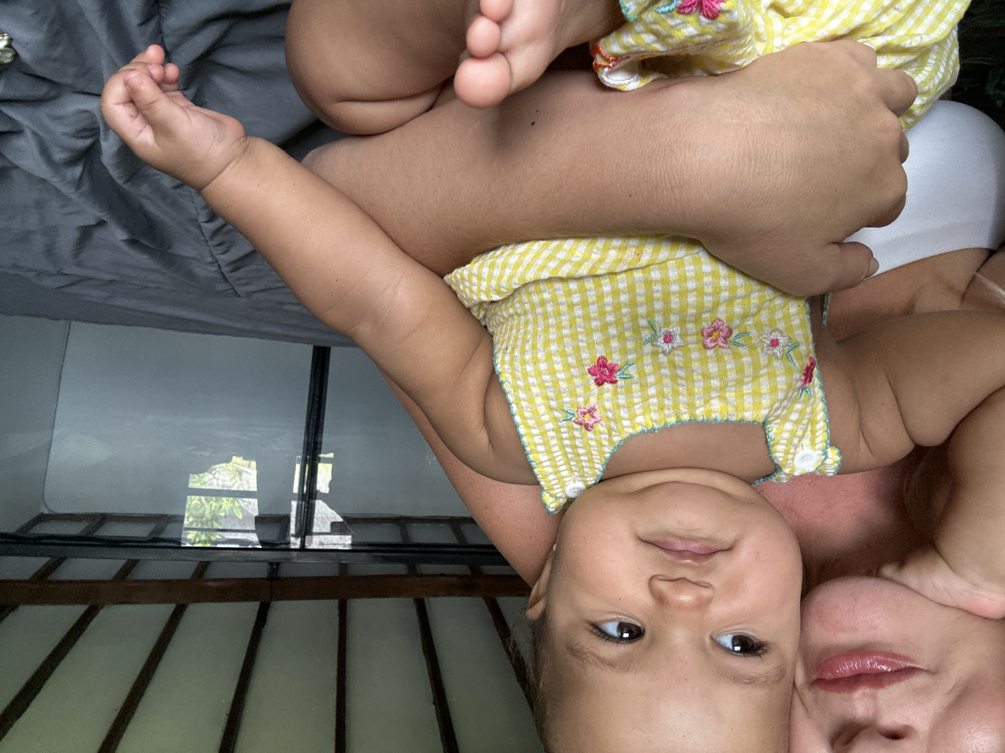 The image shows a close-up view of a woman and a baby, likely in a bedroom. The woman's face is partially visible on the left, with her lips and chin in focus. She has light skin and appears to be leaning her head on her hand. The baby, positioned in the center, has darker skin and is looking to the left with a pensive expression. The baby is wearing a yellow gingham dress with embroidered flowers. The background suggests an indoor setting with a bed covered in a gray comforter. Above, wooden beams and white ceiling panels are visible, and a window or glass pane reflects an outdoor scene with greenery, possibly indicating the location is in North Kuta, Indonesia, known for its tropical environment. The overall lighting suggests it is daytime.