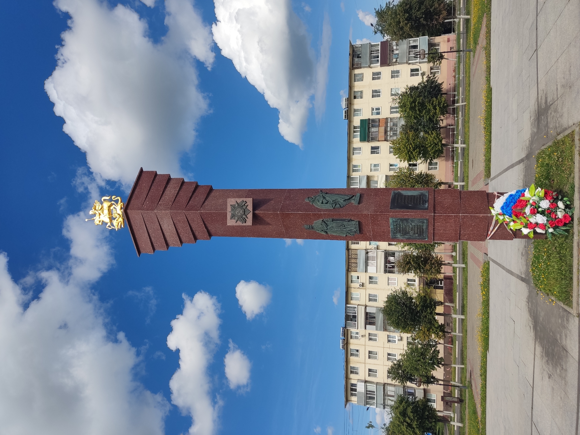 A tall, red-granite monument with a gold statue of a knight on horseback at its peak stands in a paved plaza. The monument is adorned with bronze plaques featuring historical imagery and inscriptions. To the left of the monument, a wreath of red, white, and blue flowers rests on the ground. Behind the monument, a light-colored, multi-story apartment building with numerous windows and balconies is visible. A row of trees with white trunks lines a road in front of the building, separating it from the plaza. The sky above is a vibrant blue with scattered white clouds. The scene appears to be an outdoor public space in a city.