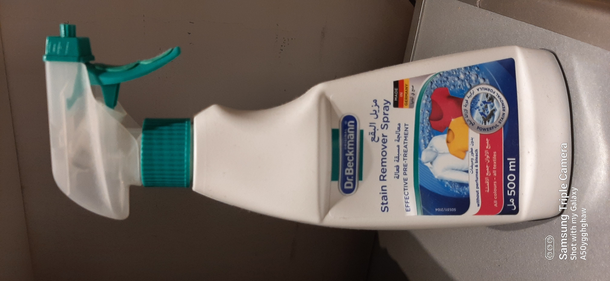 This is a close-up, eye-level, indoor photograph of a Dr. Beckmann Stain Remover Spray bottle. The bottle is white with a teal spray nozzle and cap. The label is primarily white and blue, with text in English and Arabic. The English text reads "Dr. Beckmann," "Stain Remover Spray," "EFFECTIVE PRE-TREATMENT," "without perfumes & bleach," "All colours - all textiles," and "500 ml." There is also a circular graphic depicting shirts being cleaned. A small flag indicates "MADE IN GERMANY." The background is a blurred, neutral gray. The bottom of the image includes a watermark that reads "Samsung Triple Camera" and "Shot with my Galaxy A50yggghaw." There is no indication that this image was taken in Dubai, United Arab Emirates.