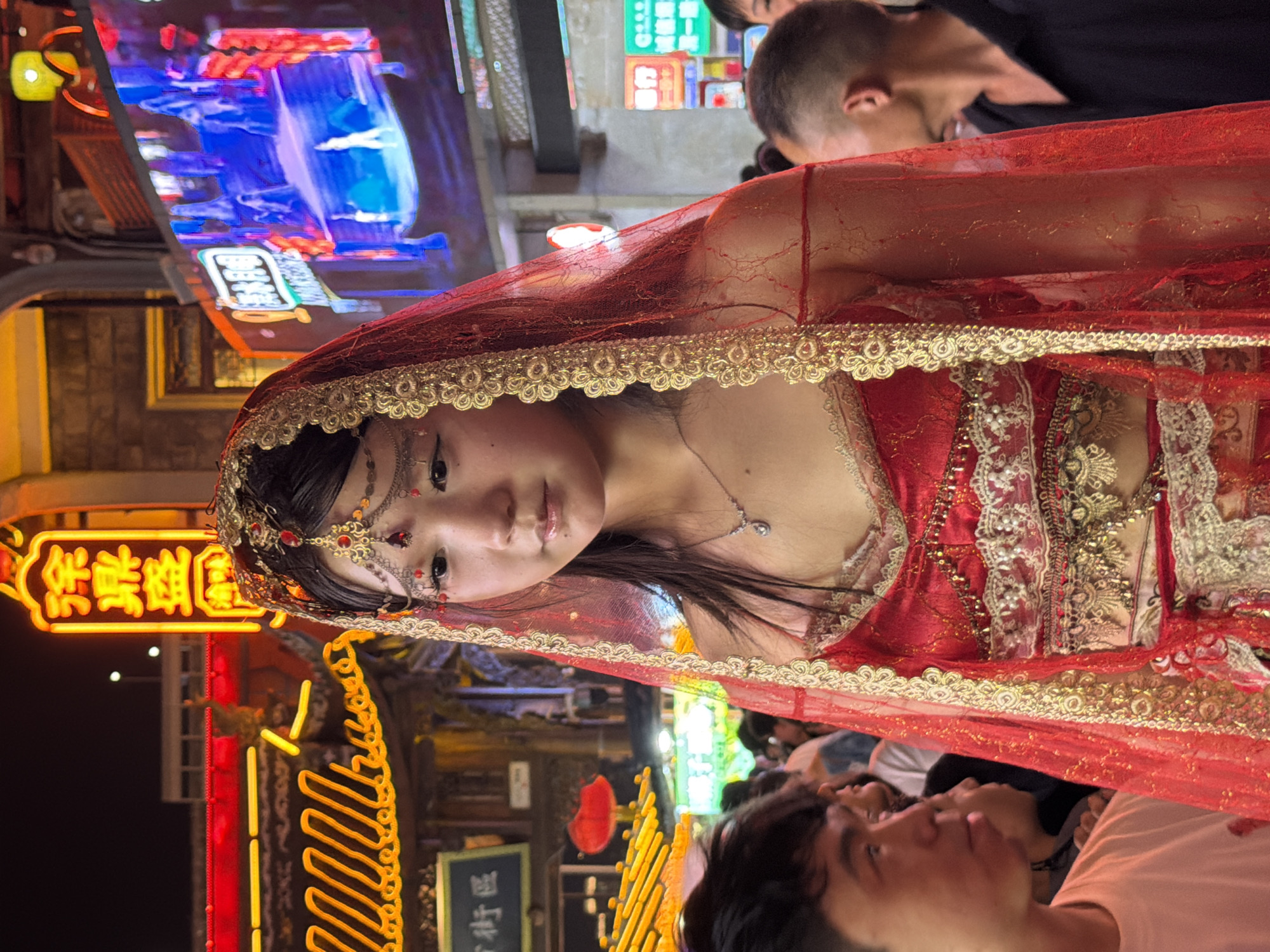 In Yuzhong District, China, a young East Asian woman, adorned in a red and gold traditional Indian-style bridal outfit and veil, is captured in a medium shot, facing the viewer with a neutral expression. Her makeup is noticeable, with pronounced eyeliner and eyeshadow. The background is a bustling night scene with brightly lit neon signs and displays, characteristic of a commercial or entertainment district. In the foreground, to her left, the shoulder and head of another individual are visible, wearing a white shirt. To her right, a man in a black collared shirt is partially visible in profile. The overall impression is of a cultural event or performance taking place in a vibrant urban setting.
