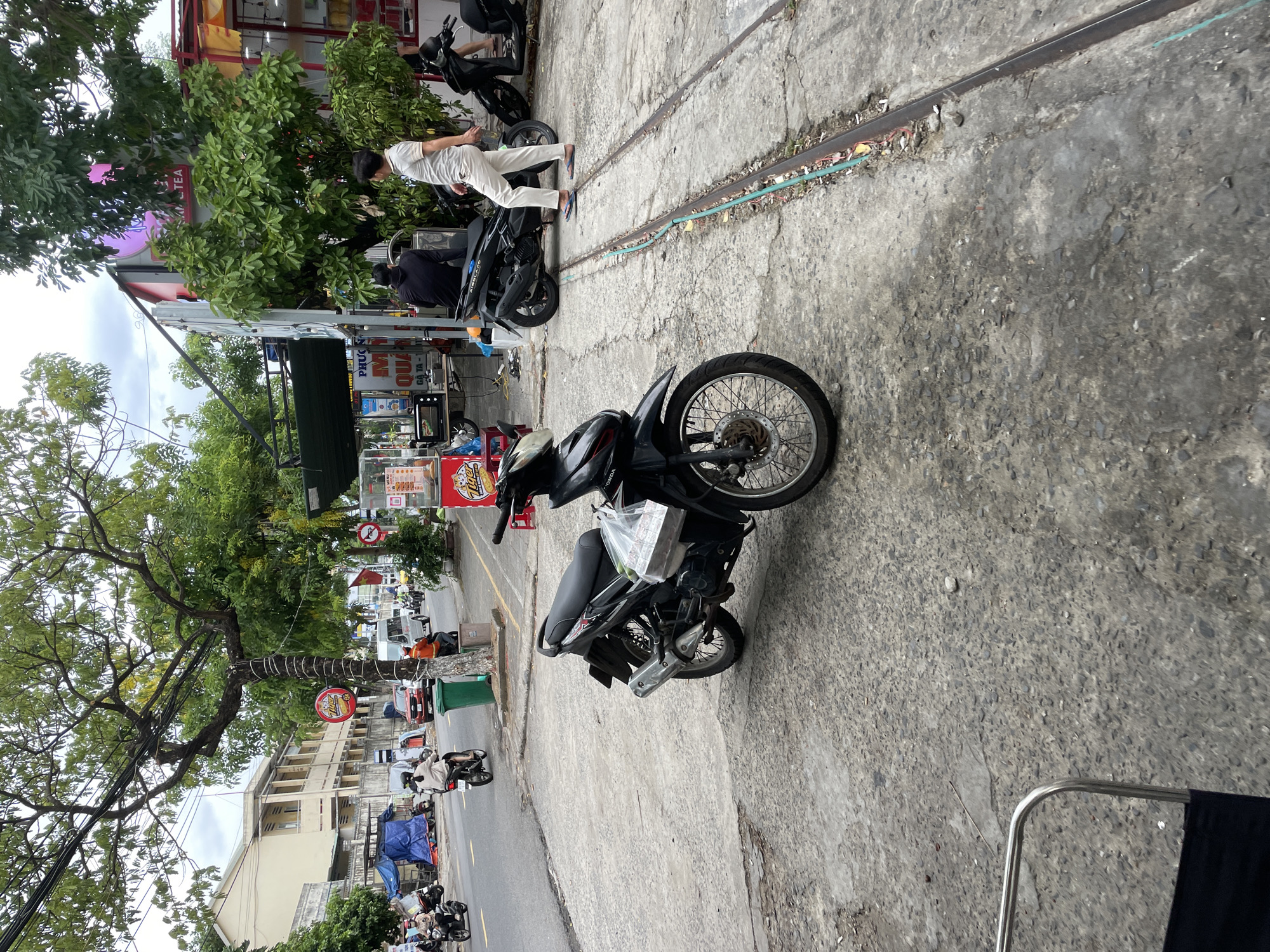 A black Honda motorcycle is parked on a concrete sidewalk in front of a building with red and white signage. A person in a white shirt and light-colored pants walks past the motorcycle towards the right side of the frame. Another black motorcycle is parked behind the first one, and a person in a dark jacket is seated on it. The street is visible in the background with several other motorcycles and a white van. A tree with green leaves shades the area. The ground is cracked concrete with what appear to be old train tracks running through it. The overall scene suggests a busy street or market area in a city.