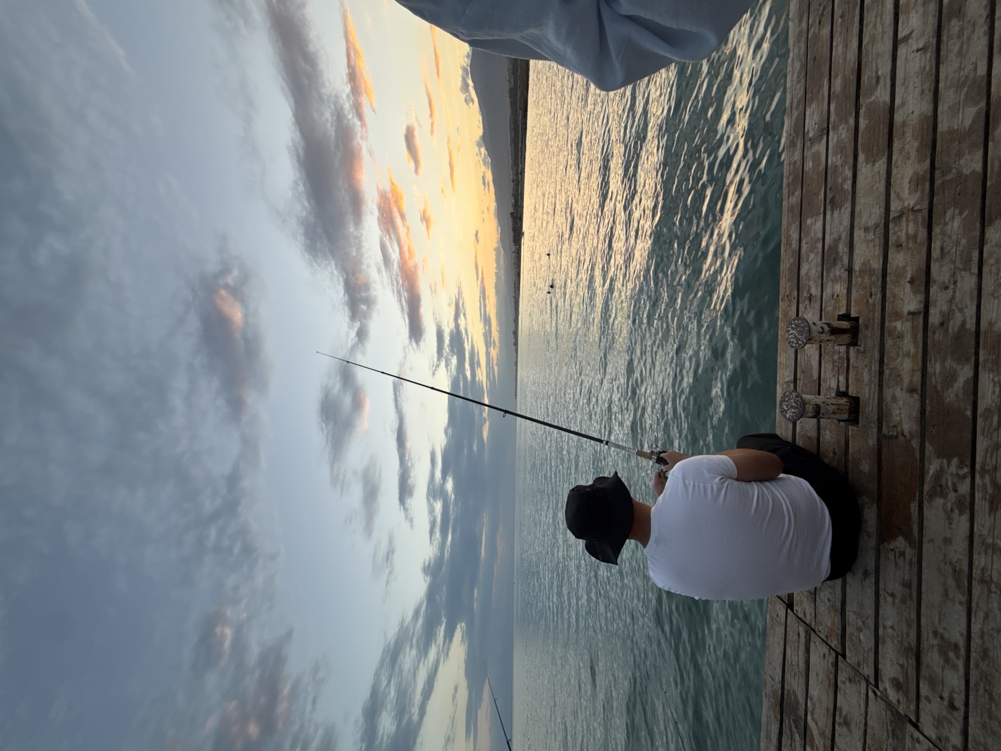 A person wearing a white t-shirt and a black bucket hat is sitting on a wooden pier, fishing. They are facing away from the camera, looking out at the calm water. The sun is setting, casting a warm glow on the sky and water. In the distance, there are mountains and a few other people swimming. The pier is made of weathered wooden planks.