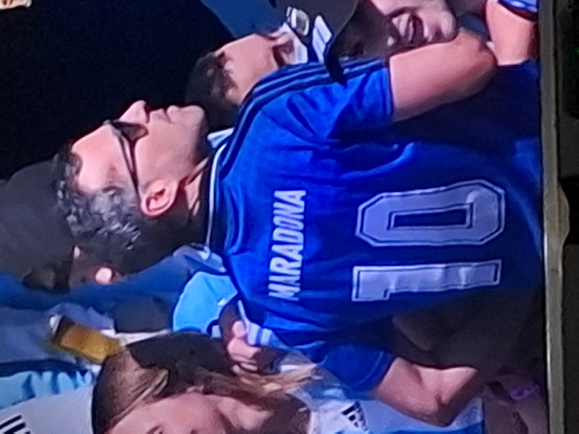 The image shows a close-up of a person wearing a blue soccer jersey with "MARADONA" and the number "10" on the back. The jersey appears to be a replica of the Argentinian national team jersey. The person is wearing sunglasses and is partially obscured by other people. The image is taken from a slight distance and appears to be a candid shot, possibly at a sporting event. There is no visible indication of the specific location within Almaty, Kazakhstan. The overall lighting is dim, suggesting an indoor or nighttime setting.