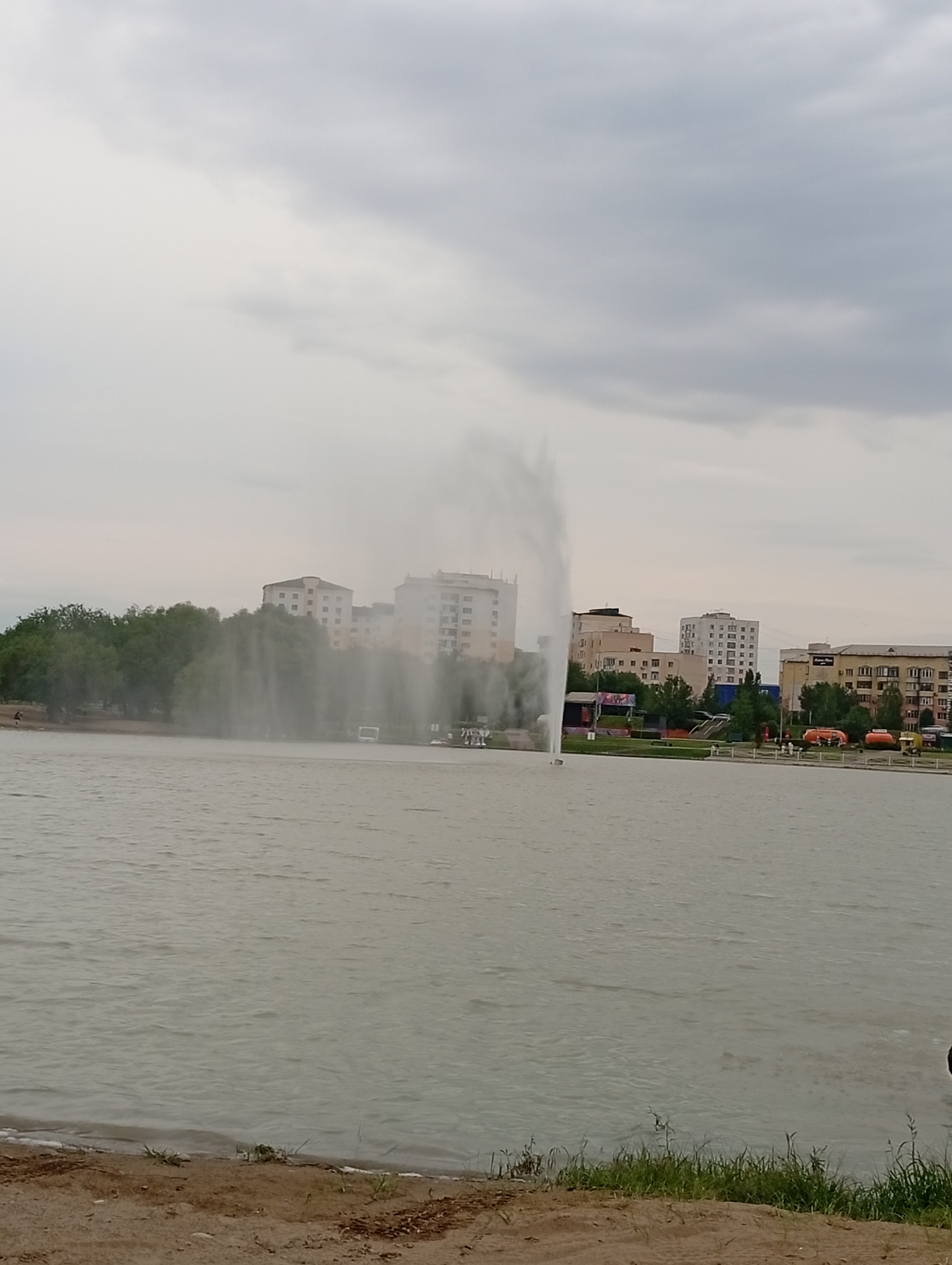 The image depicts a wide, calm body of water in front of a cityscape under a cloudy sky. A large fountain sprays water high into the air in the middle of the water. In the background, several multi-story buildings, some appearing to be residential complexes, are visible behind a line of lush green trees. Various vehicles, including what appear to be orange industrial trucks, are parked along the waterfront. The shore in the foreground is a sandy beach with sparse patches of grass. A few indistinct figures can be seen on the distant shore, suggesting it's a public area. The overall impression is a serene, possibly urban, park setting on an overcast day, with the fountain serving as a central point of interest. No visible text is present in the image.