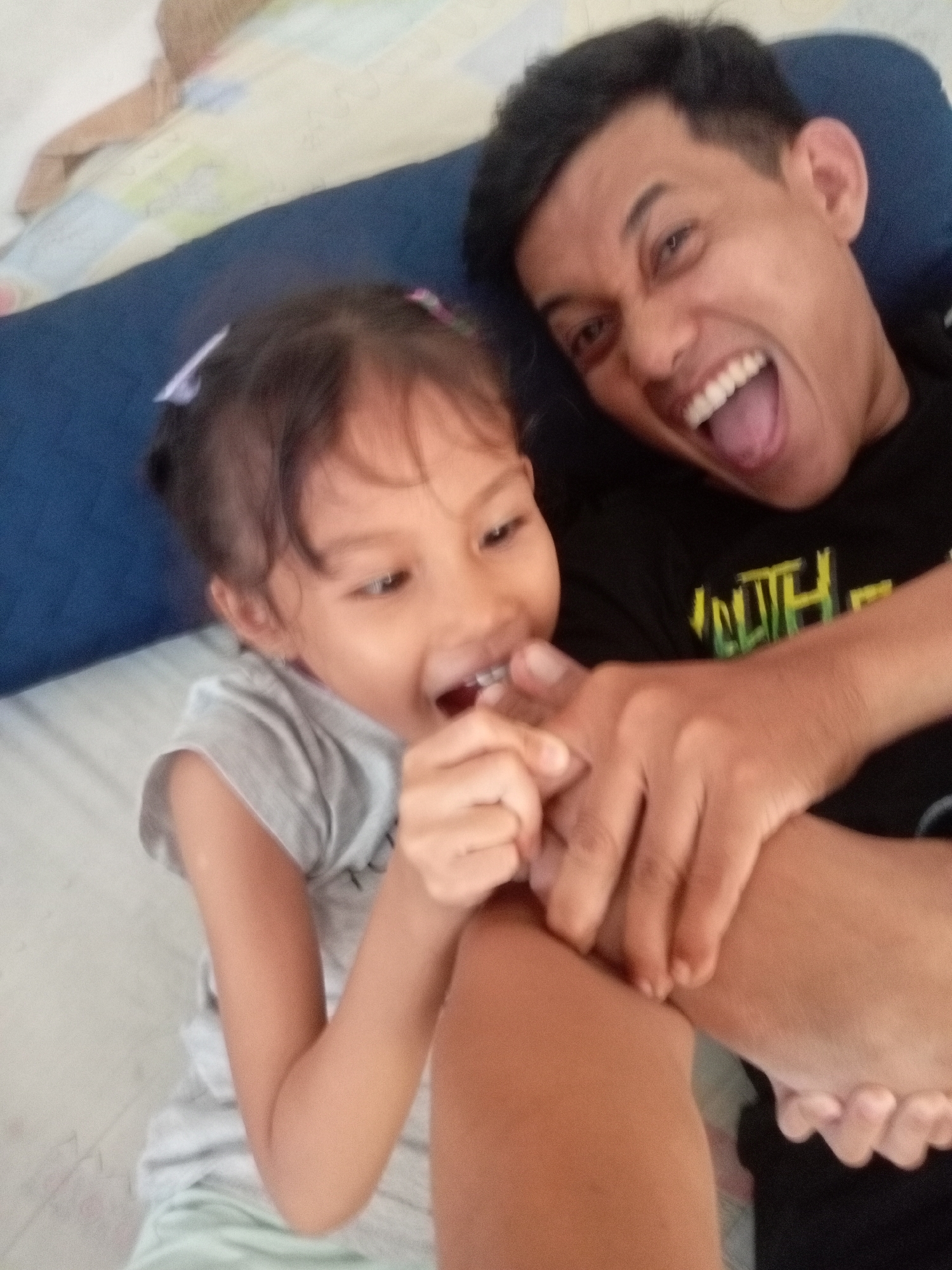 In Batam, Indonesia, a father and daughter are captured in a close-up selfie. The father, an adult male with short dark hair and a black t-shirt, is on the right side of the frame, his mouth wide open in a laugh, revealing his teeth and tongue. His right arm is extended, and his hand is touching his daughter's face. The daughter, a young girl with dark hair adorned with a white and pink hair clip, is on the left. She is wearing a grey t-shirt and is looking towards the camera with her mouth open, touching something near her mouth with her right hand. They are lying on a bed with a blue bolster pillow behind them and a patterned sheet beneath. The overall atmosphere is one of playful interaction and affection.