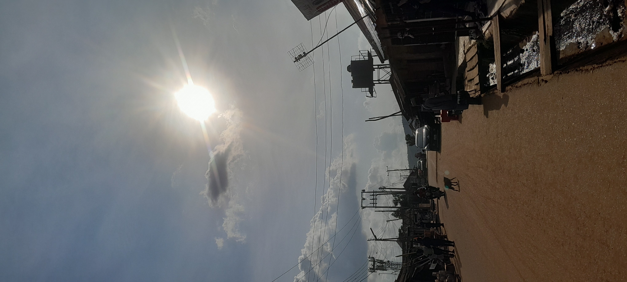 In a street scene in Abuja, Nigeria, the bright sun shines from a hazy sky, casting rays across the image. The unpaved road is dusty brown, with a slight sheen from water in a drain on the right side of the frame. Wooden planks bridge the drain. On the left side of the road, several people are visible, some walking and others standing near stalls or shops. A motorcycle is being ridden down the street, with a passenger behind the rider. A dark-colored car is parked further down the road. A dog is walking in the middle of the road. Buildings line both sides of the street, appearing to be a mix of residential and commercial structures, some with corrugated iron roofs. Electrical poles with wires crisscross the sky. In the background, lush green hills are visible. A water tank on a metal structure stands above some of the buildings on the right.