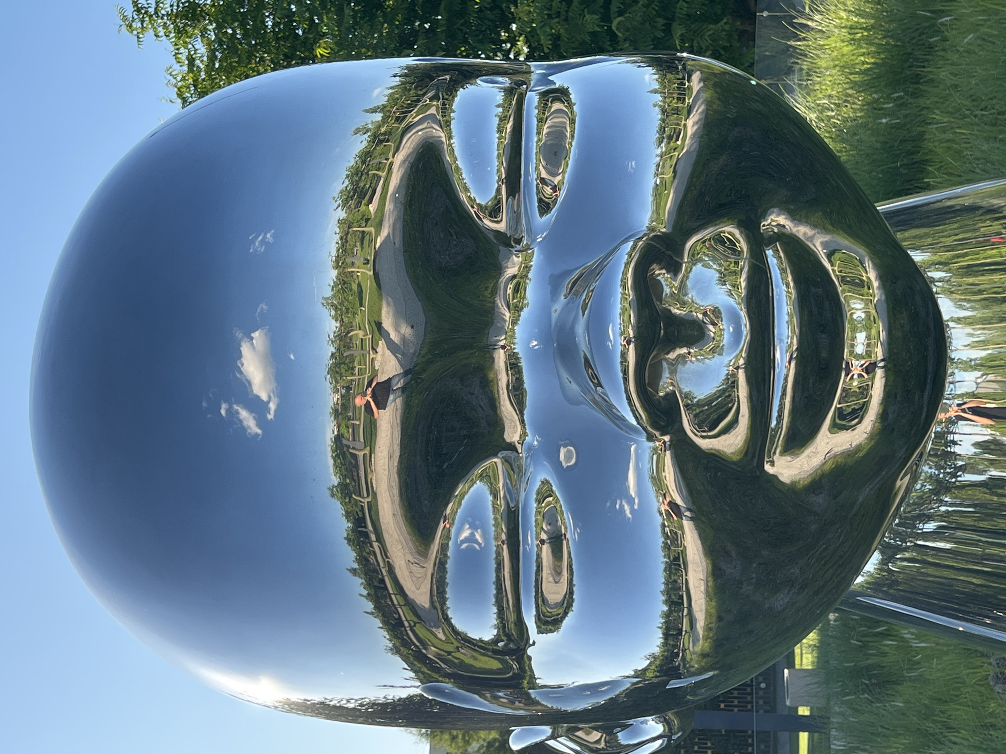 The media file features a large, highly polished, chrome-like metallic sculpture of a human head or face, positioned outdoors under a bright blue sky. The sculpture's surface is extremely reflective, acting like a distorted mirror and showing warped reflections of its surroundings.
The reflections on the sculpture's surface reveal a park-like setting with lush green trees, grass, and a paved path. The upper part of the sculpture primarily reflects the clear blue sky with a few white clouds. On the "face" area, the distorted reflections of the path and foliage create abstract shapes that resemble eyes, a nose, and a mouth, giving the impression of facial features.
Several human figures are visible in the reflections, stretched and distorted by the sculpture's curved surface. They appear to be walking on the path or standing nearby, observing the artwork. One figure is seen wearing a dark top on the right side of the reflected face, and another on the left.
The scene depicts bright daylight, suggesting a sunny morning or afternoon. The weather is clear, as indicated by the vivid blue sky and sharp reflections. The overall setting is an outdoor art installation within a well-maintained green space, likely a park, in Krasnodar, Russia. There is no visible text in the image. The unique reflectivity and distortion transform the landscape into an abstract visual experience on the sculpture's surface.