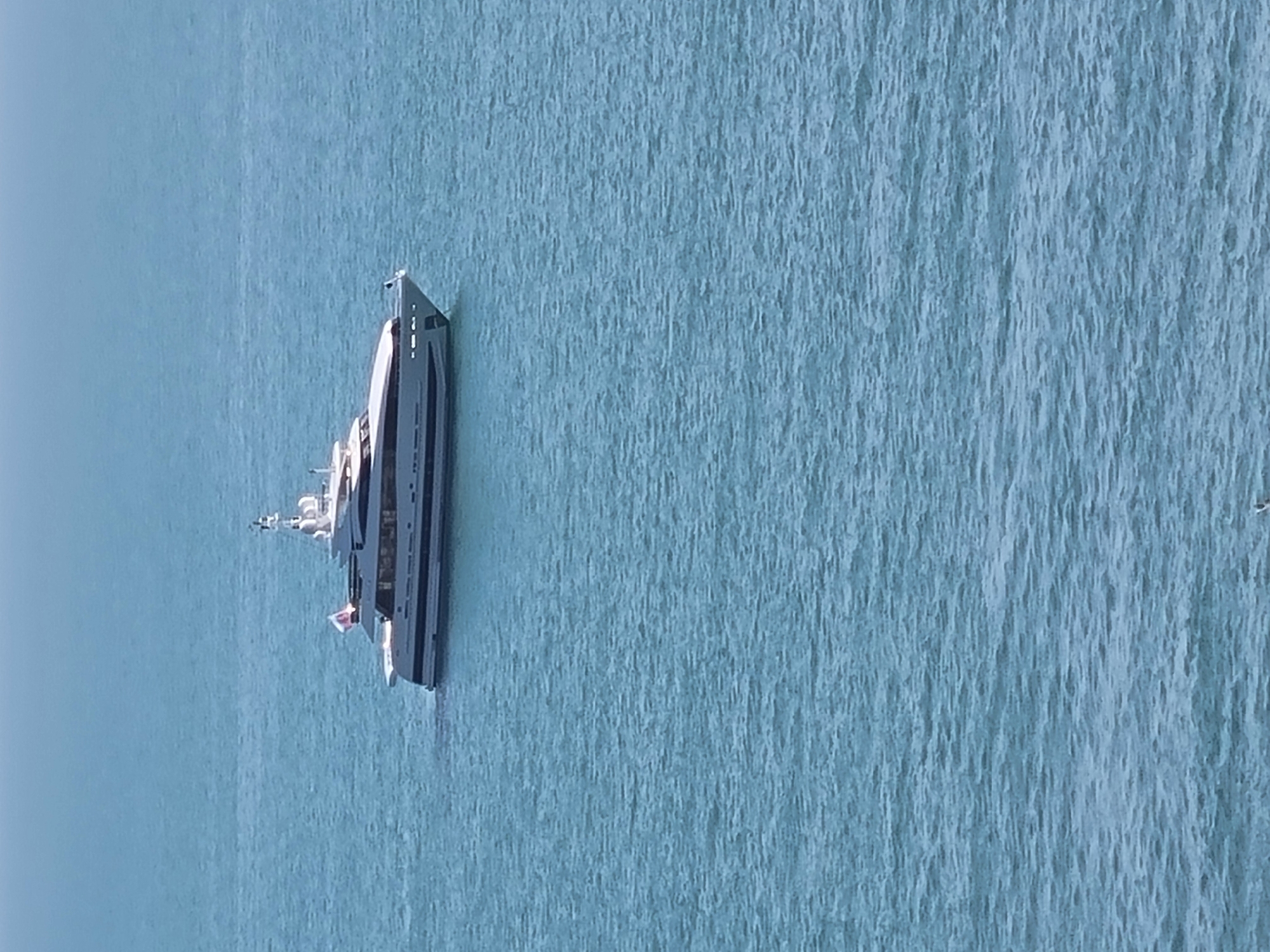 A luxury yacht floats on turquoise water. The location is not explicitly discernible as Sochi, Russia, from the image. The yacht is grey and white, with a multi-tiered structure and a flag. The water texture appears rippled and slightly choppy. The sky is a light blue. The image is shot from an elevated viewpoint.