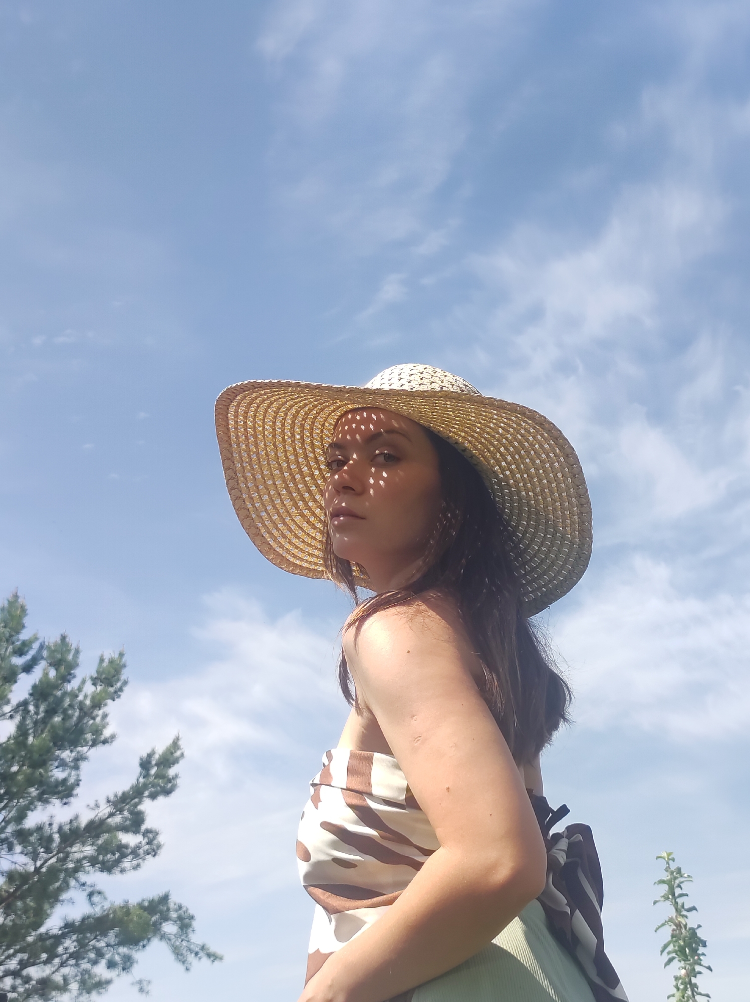 A young woman stands outdoors on a sunny day, wearing a wide-brimmed straw hat that casts patterned shadows on her face. She is wearing a brown and white patterned top and a green skirt. A striped bag hangs from her shoulder. Behind her, there's a blue sky with light, wispy clouds, a pine tree, and a small leafy plant. The location is outside and appears to be a sunny day. The city location of Фоки, Russia is unsupported by the image.