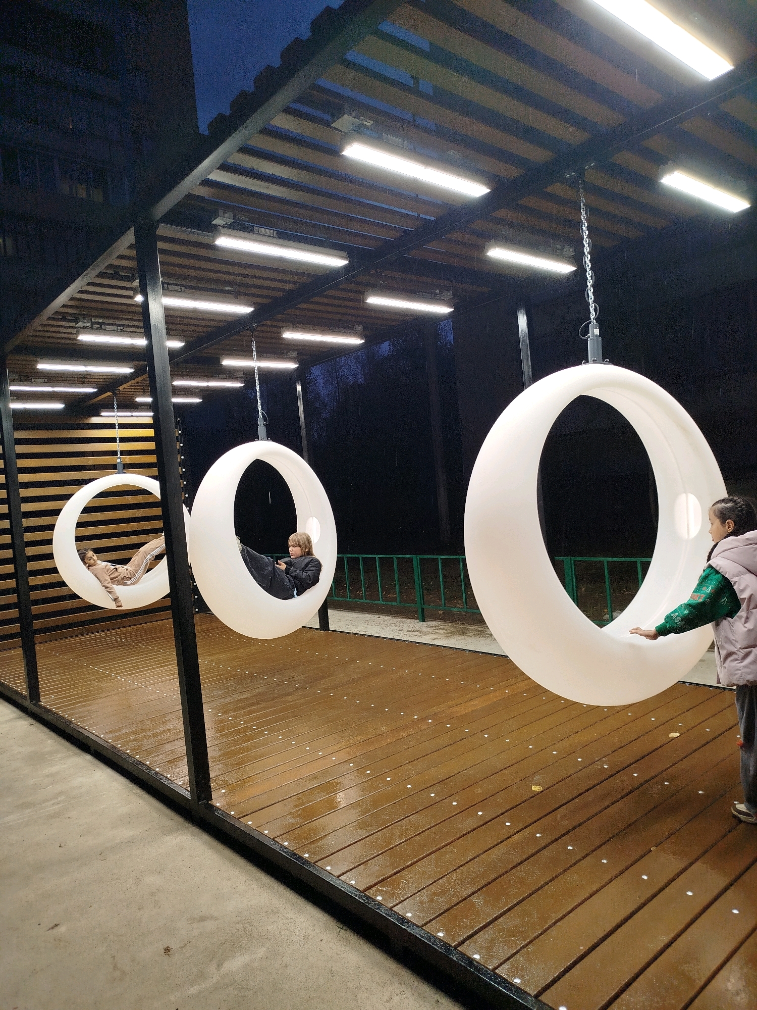 In the city of Unknown, Unknown, an outdoor area features a modern, covered structure with a slatted wooden roof and numerous fluorescent lights. The structure has dark metal beams supporting it. Three large, white, circular, illuminated swings are suspended from the ceiling by chains. The ground within the structure is a wooden deck, wet from rain, with small, round lights embedded in it. To the left, a person lies relaxed in one of the swings, dressed in a light brown tracksuit. In the middle, another person, a child, sits in a swing, looking at a phone, wearing a dark outfit. To the right, a girl wearing a light pink puffer vest over a green sweater and dark pants stands next to a swing, reaching out to touch it. The background is dark, with trees and a fence visible beyond the structure. A dark building is visible to the far left, and the sky is a deep blue, indicating it is evening or night.