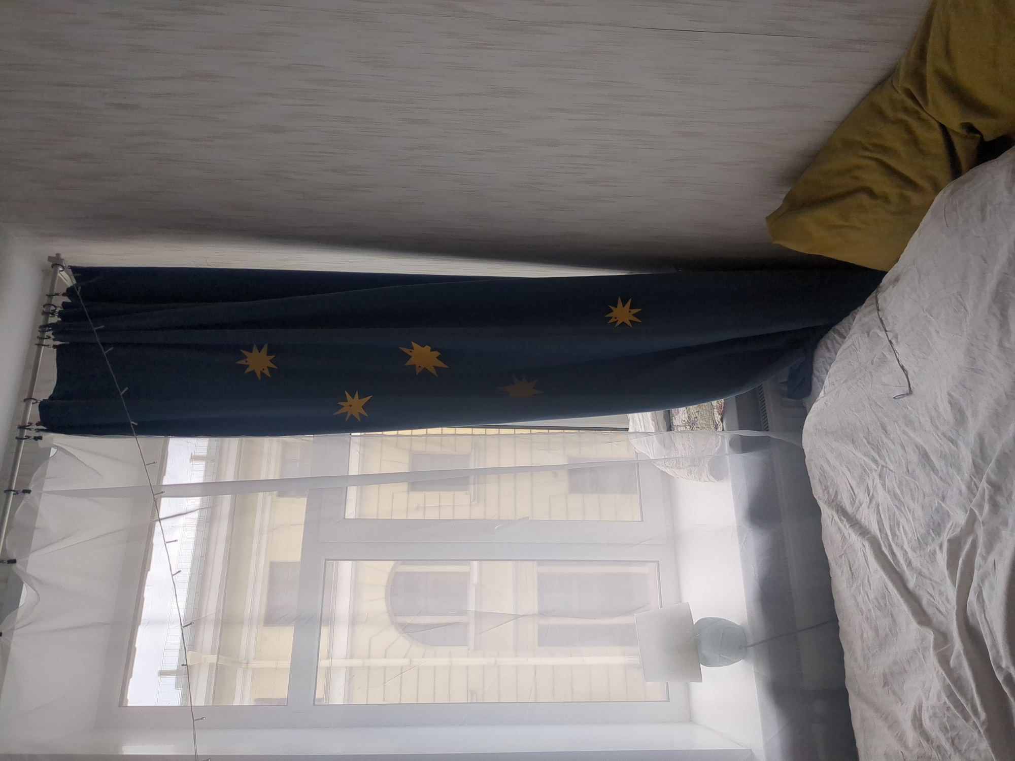 A low-angle, slightly tilted, full-color image depicts a bedroom interior in an apartment, potentially in Saint Petersburg, Russia. The primary focus is a window covered by two layers of curtains. The outer layer is a dark blue curtain with scattered golden star patterns. The inner layer consists of sheer, white curtains, allowing light to filter through. Beyond the window, the exterior view shows a building with beige or light yellow facade, likely residential, and is characterized by rectangular windows and an arched architectural detail. To the right of the window, a bed is partially visible, covered with a rumpled white sheet and a yellow pillow. A green lamp sits on a table in front of the window. The ceiling is white and appears to have a textured or wooden-planked surface. A string of fairy lights is visible hanging in front of the window. The overall scene suggests a cozy, lived-in space.