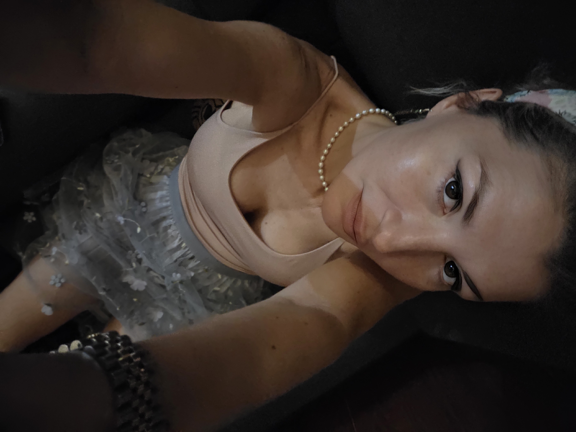 A close-up, overhead shot shows a woman from the chest up. She is looking directly at the camera with a neutral expression. She has on a light pink tank top and a sheer, light gray skirt with small white floral appliques. A pearl necklace is around her neck, and she is wearing a beaded bracelet on her right wrist. Her hair is pulled back and she has a colorful bandana in it. The background is dark and out of focus, but it appears to be an indoor setting. There is no indication of location in North Kuta, Indonesia.