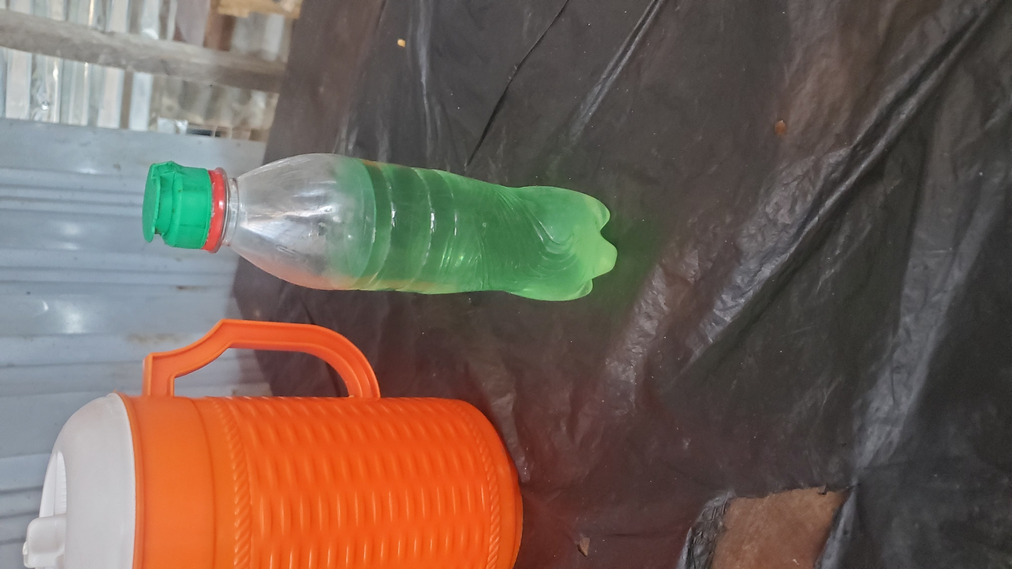 In Jalingo, Nigeria, on a black plastic tablecloth, an orange thermos with a white lid and handle sits next to a clear plastic bottle filled with green liquid. The thermos has a textured pattern and a large handle. The bottle has a green flip-top lid with a red seal. The background shows a corrugated metal wall and some wooden structures.