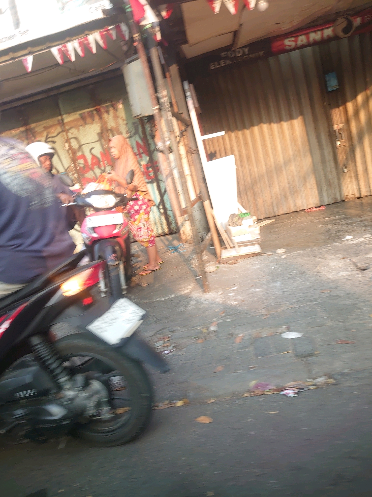 The image depicts a street scene in South Jakarta, Indonesia, with a focus on a red and black scooter in the foreground, viewed from the left rear. A person wearing a dark blue shirt is partially visible on the scooter, with a white helmet. Behind this scooter, another scooter, primarily red, is parked. A person wearing a peach-colored hijab and a patterned dress is standing next to the red scooter, holding a smartphone. The background reveals a corrugated metal wall of a building, likely a shop, with the signage "FODY ELEKTRONIK" and "SANKI" visible. Red and white triangular flags are strung above the building's entrance. The ground is asphalt, with scattered debris and fallen leaves, indicating an outdoor urban environment. The lighting suggests it is daytime, with sunlight casting shadows. The overall impression is of a typical street scene in a commercial area.