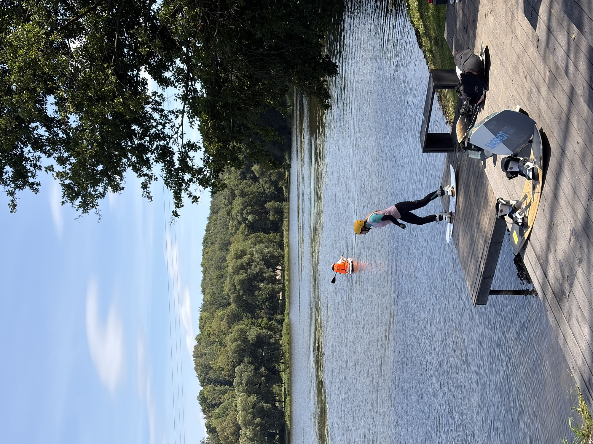 A person in a yellow helmet and pink vest is standing on a wakeboard on a wooden dock, facing away from the camera and towards a lake. Another person wearing an orange life vest is paddleboarding in the distance on the lake. The lake is surrounded by a dense forest. The sky is blue with scattered clouds. Several wakeboards and bindings are laid out on the dock. A black bench is positioned on the dock behind the person on the wakeboard. The scene is set in Astrashitskagaradokski rural council, Belarus.