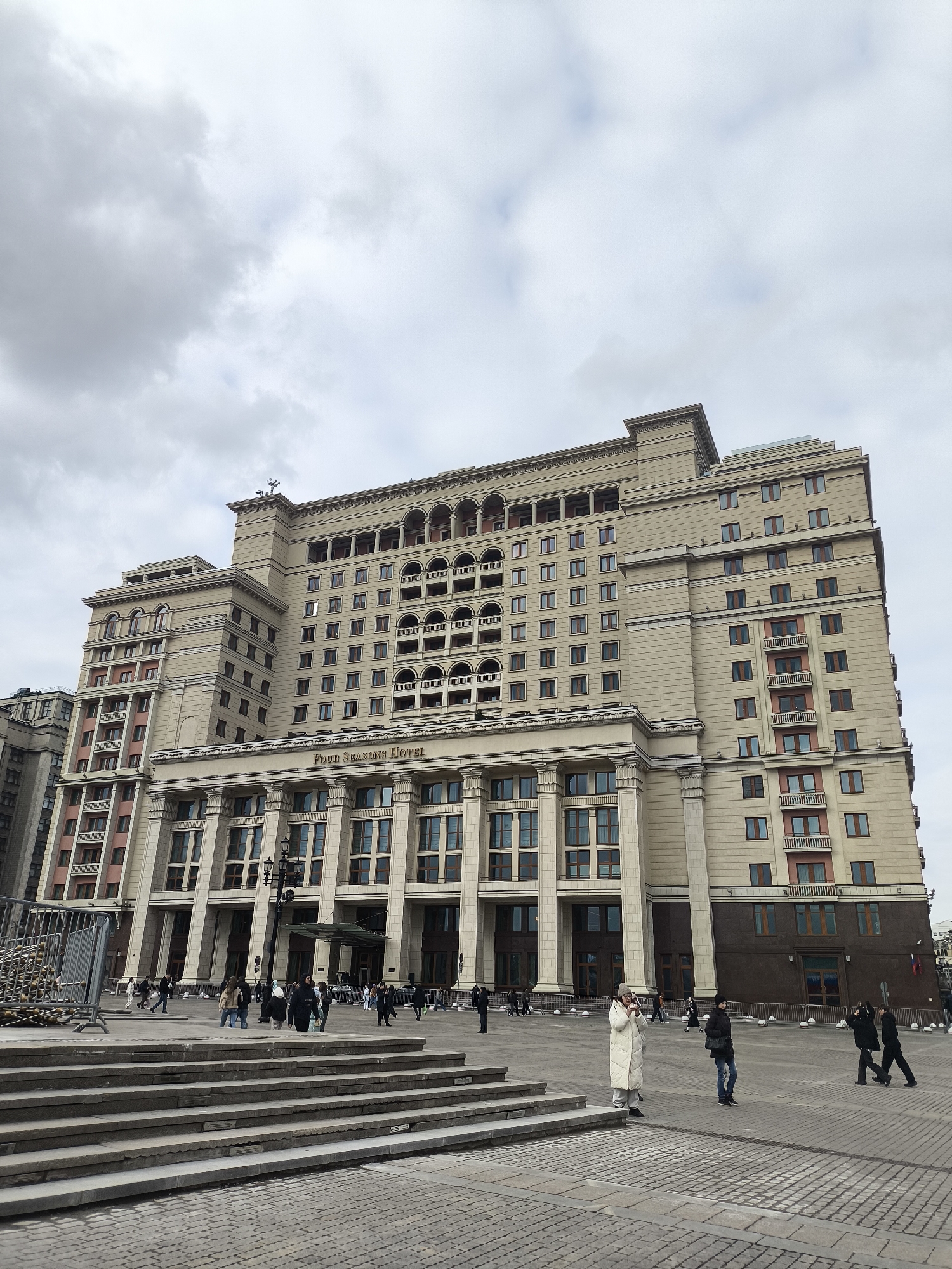 A wide-angle, eye-level shot captures the grand facade of the Four Seasons Hotel in Moscow, Russia, under an overcast sky. The imposing building dominates the frame, showcasing ornate classical architecture with numerous windows, columns, arches, and decorative cornices, rendered in light beige or off-white stone with darker accents. The hotel's name, "Four Seasons Hotel," is prominently displayed above the main entrance, which is flanked by large columns.
In the foreground, a wide, stone-paved public square features a set of broad, shallow steps leading up from the lower left. Scattered across the plaza and near the hotel entrance are several pedestrians, engaged in typical urban activities like walking or observing. One individual in a long white coat stands near the foreground steps, seemingly engrossed in their phone or taking a picture. Others wear casual attire, including coats and jackets, suggesting cool weather.
To the left, stacked metal barriers or fencing are visible, possibly indicating ongoing work or event preparations. Street lights are present near the hotel's entrance. The overall scene depicts a busy, public space in a major city, characterized by monumental architecture and diffused daylight from a cloudy sky.