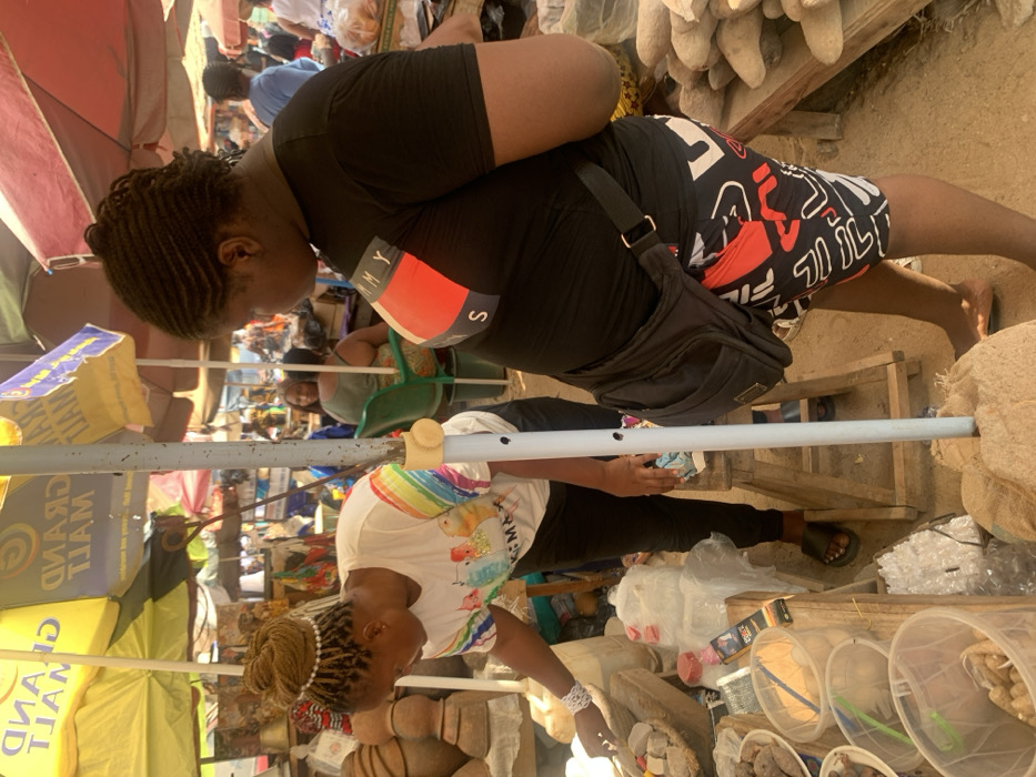 The scene depicts an outdoor market in Otuocha, Nigeria, under bright daylight, partially shaded by multiple large, colorful market umbrellas, including a yellow "GRAND MALL" branded umbrella. In the foreground, two women are present. The woman on the left, with blonde braided hair adorned with a pearl headband, wears a white t-shirt with a rainbow graphic and dark pants, and black sandals. She is leaning forward, actively examining or arranging various goods displayed on a wooden stall, which includes natural products like roots, powders in clear containers, and packaged items. To the right, another woman with dark braided hair (cornrows) wears a black t-shirt with a red, white, and blue "TOMMY S" logo, black, white, and red patterned shorts, and carries a black crossbody bag. She stands barefoot, facing generally towards the left woman and the stall. A light-colored pole, potentially supporting an umbrella or stall, stands centrally, anchored in a burlap sack. The stall to the right displays large, light-colored root vegetables. In the background, other market stalls, people, and a green plastic chair with a seated individual are visible, indicating a busy market environment with a dirt ground.