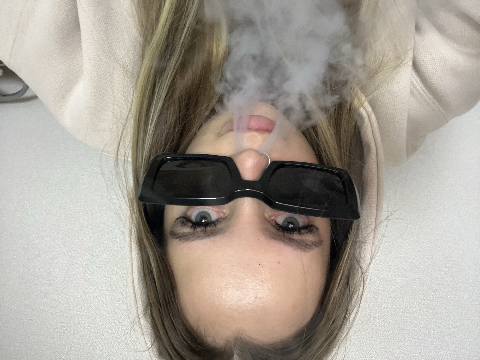 A close-up, rotated portrait depicts a person with their head positioned at the bottom of the frame, exhaling a plume of white smoke or vapor that rises upwards. The individual has long, light brown hair with blonde streaks, framing their face. They are wearing modern, rectangular black sunglasses, which are pushed up slightly to reveal striking blue-green eyes and prominent eyelashes looking directly into the camera. A small silver hoop nose ring is visible on the left nostril. The person is dressed in a light-colored (possibly beige or cream) hooded sweatshirt, with parts of the hood visible around their face. The background is a plain, light-colored surface, likely a wall or ceiling, offering no additional context. The overall impression is neutral, with a cool or edgy aesthetic due to the sunglasses and smoke. No text is visible in the image. This scene takes place in Krasnodar, Russia.