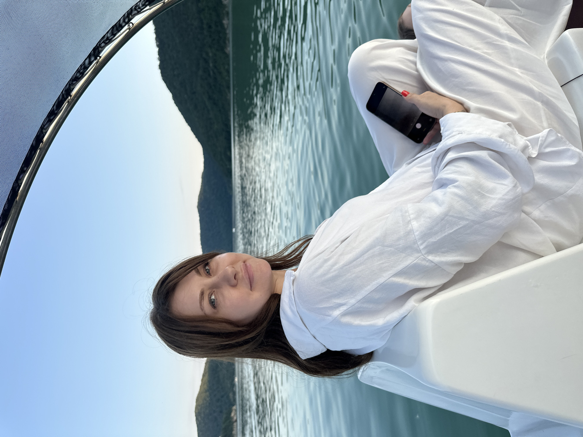 A woman with long, brown hair and blue eyes is captured in a vertical, outdoor portrait. She is wearing a white, oversized shirt and is seated on a white boat, holding a black smartphone in her right hand. The phone screen is dark, and the camera is facing the woman. Her fingernails are painted red. She is looking towards the camera with a slight smile.
The boat is on a body of water, likely a lake or reservoir, with calm, rippling green water. In the background, a lush green hillside or mountain range is visible, with a clear blue sky above. The image appears to be taken during the daytime, possibly late afternoon or early evening, given the soft lighting.
The setting suggests a leisurely activity, such as a boat trip or cruise, in a natural environment. The overall mood is relaxed and serene. The location context provided is Abrau-Dyurso, Russia, which is known for its wineries and lakes. There is no visible text in the image.