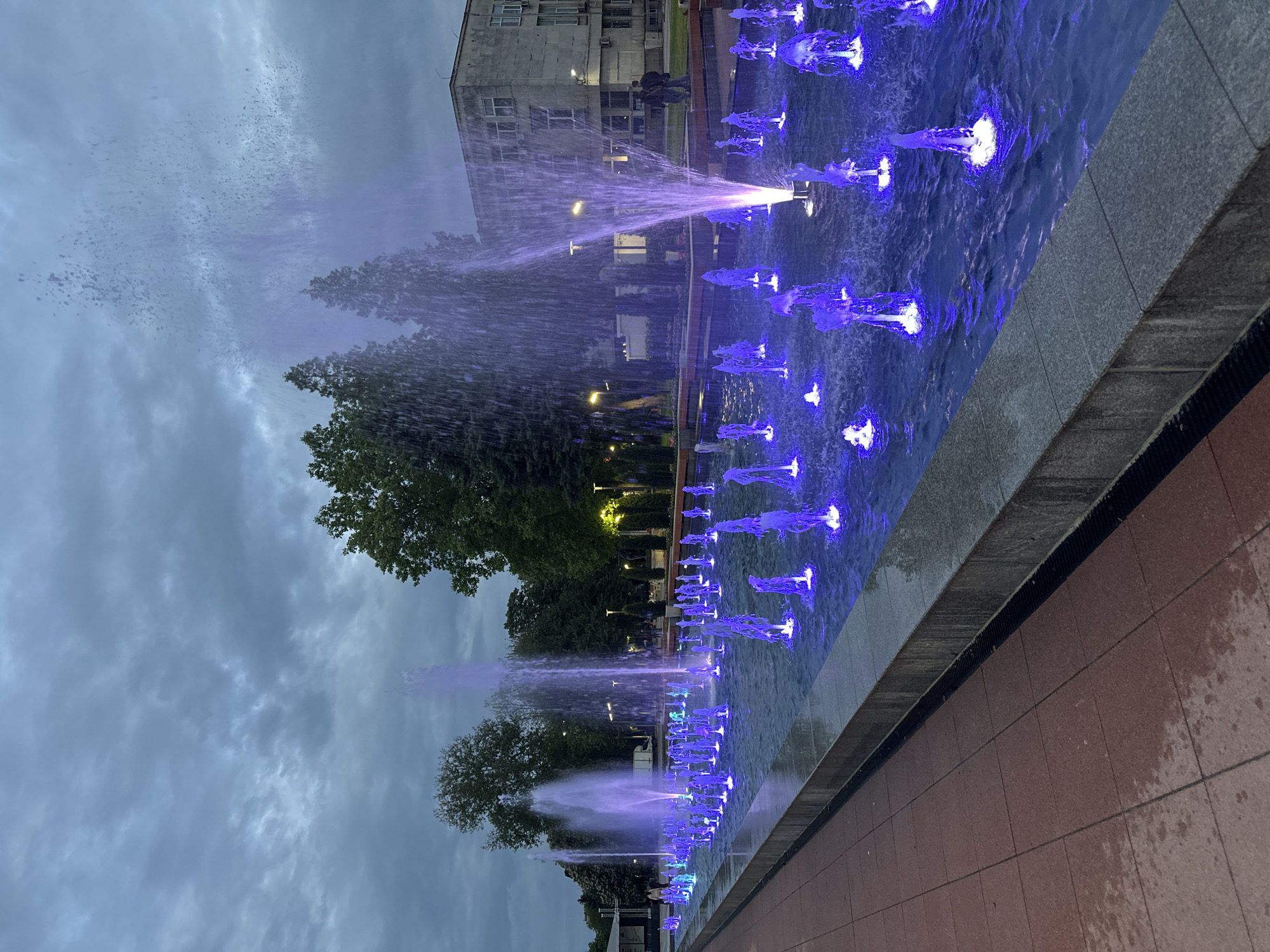 The image depicts a fountain with blue illuminated water jets, likely in a city park setting. In the background, there is a building, trees, and street lights. The foreground shows the edge of the fountain and the surrounding paved area. The sky is overcast. The photograph is taken in Montana, Bulgaria. The image is rotated 90 degrees clockwise.