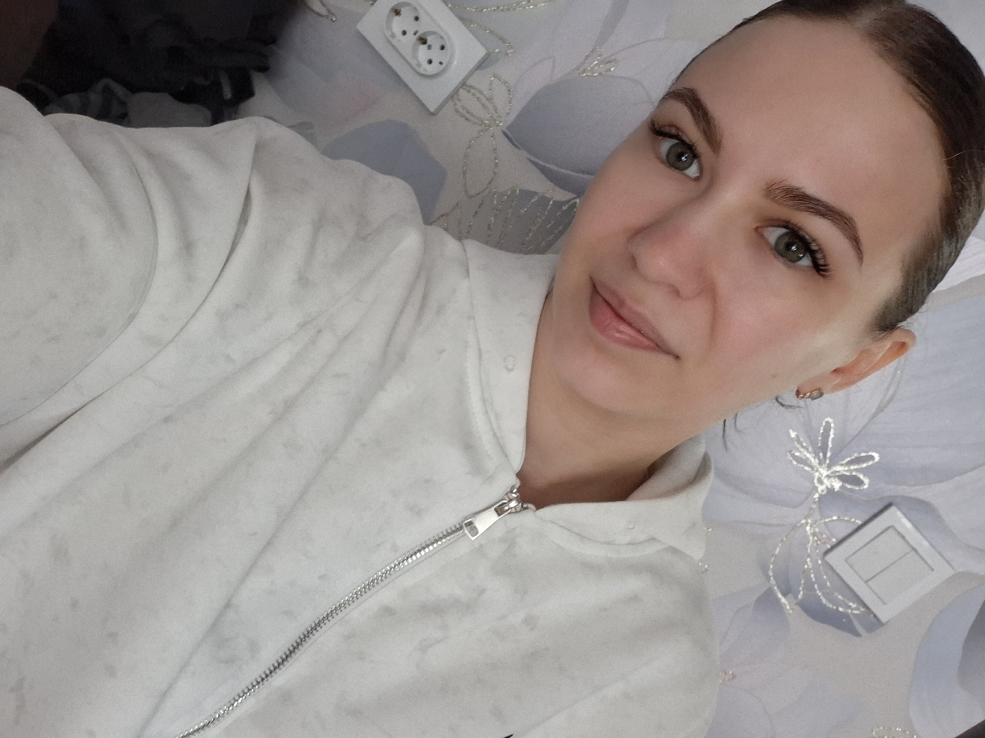 A selfie shows a woman with fair skin, brown hair pulled back, green eyes, and defined eyebrows. She wears a light-colored zipped sweatshirt and looks directly at the camera with a slight smile. The background consists of a patterned, light-colored surface, possibly bedding or a blanket, with a European-style electrical outlet and a small, square object near the lower right corner. The location is unknown and there is no proof that it is in the city of Nizhnyaya Gar', Russia.