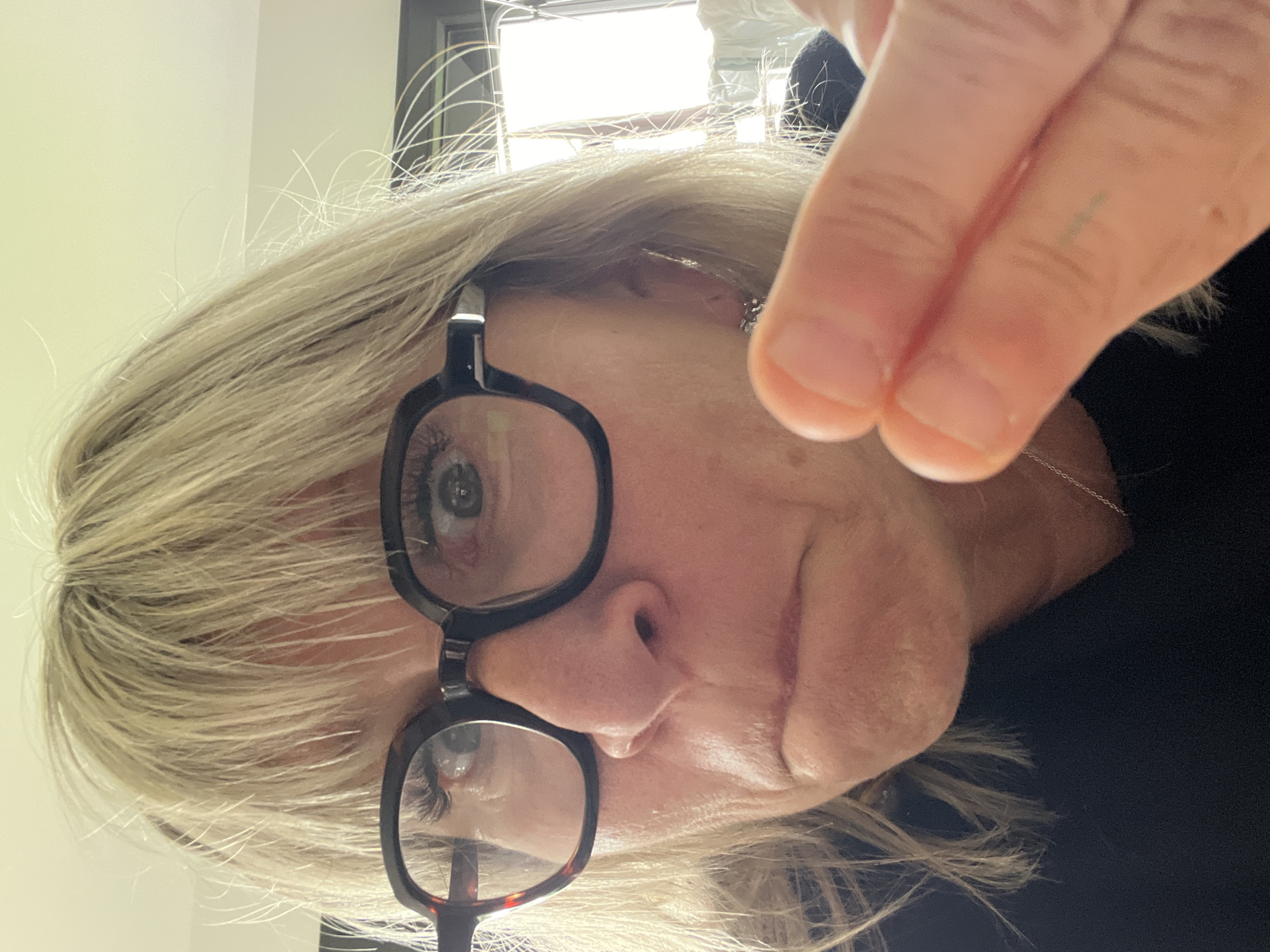 A close-up, slightly tilted, indoor photograph shows a fair-skinned woman with short, blonde hair wearing black-rimmed glasses. She is looking downwards, and her right hand is raised towards the camera. A window and part of a black garment are visible in the background. The location is not evident from the image.
