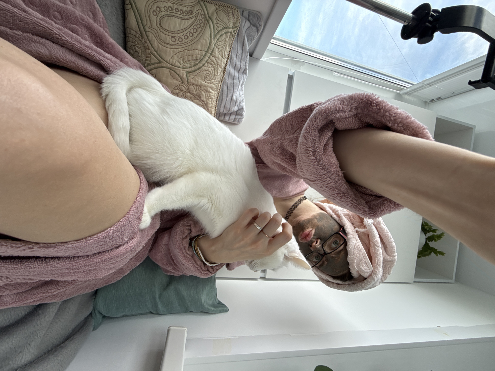 A woman with a face mask and a pink towel turban is holding a white cat. She is wearing a pink robe. The scene appears to be indoors, near a window with blue sky visible. The cat is on her lap, and she is holding it with her right arm. She is also wearing glasses and a black choker necklace.