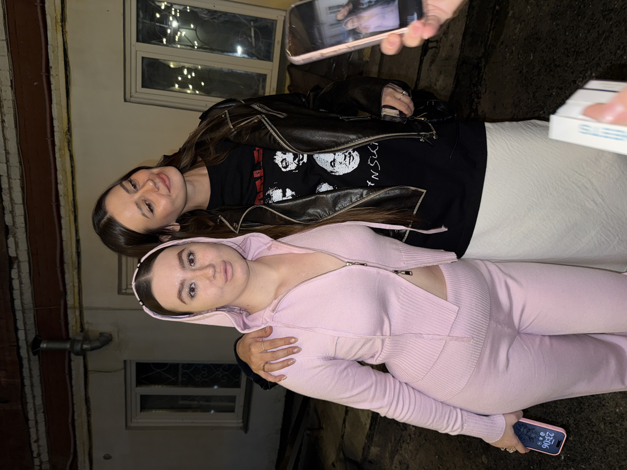 Two young women are posing for a photo in an urban alleyway in Krasnoyarsk, Russia. The woman on the bottom wears a pink knit tracksuit, while the other wears a leather jacket over a black graphic t-shirt and white pants. They are close together, with the woman in the pink tracksuit holding a smartphone. A hand holds up another smartphone in the top right corner to presumably take the photo. In the background, there are aged walls, windows with string lights, and pipes.