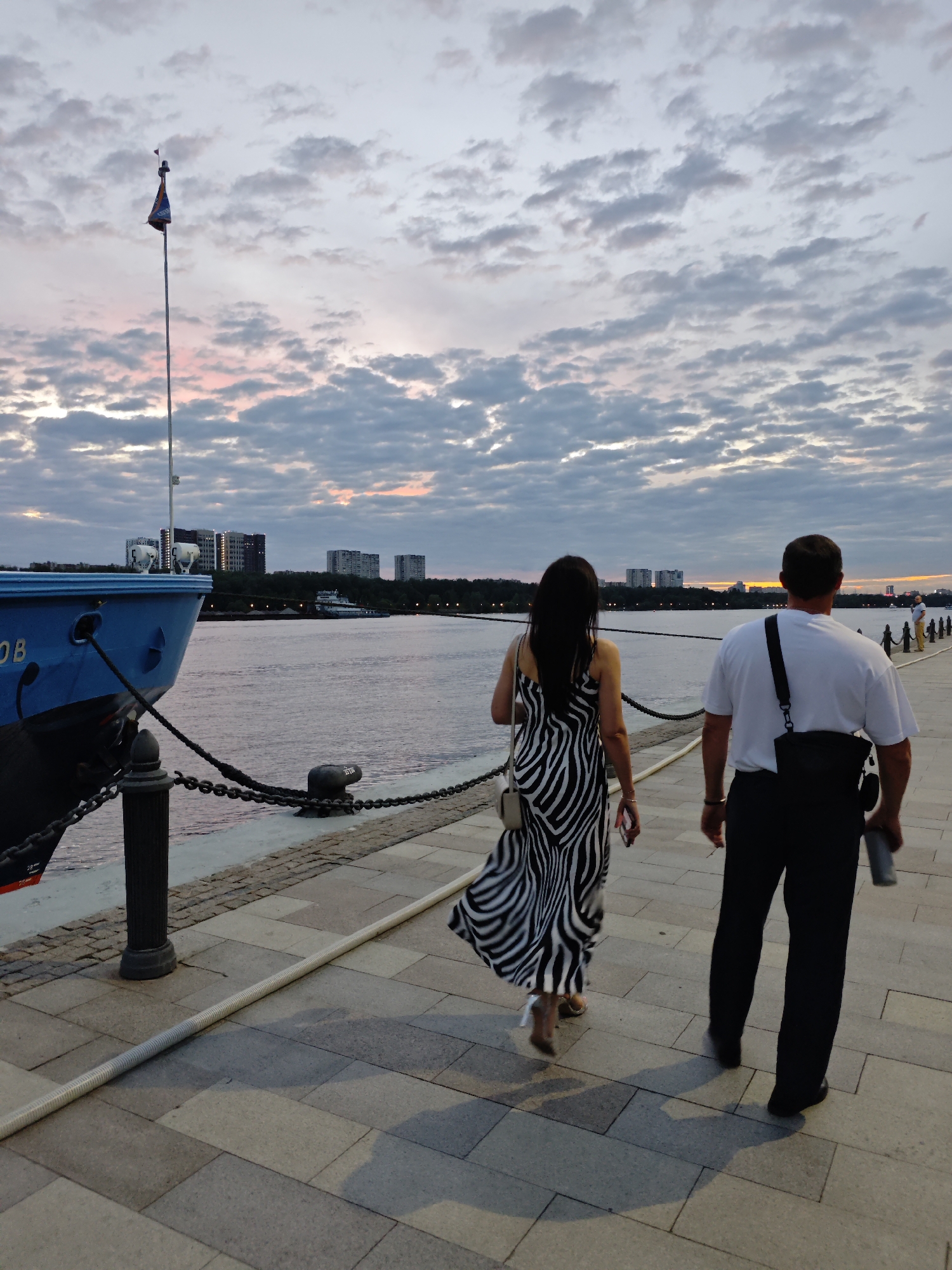 At dusk, a couple walks away from the camera on a paved embankment along a wide river, likely the Moskva River in Moscow, Russia. The woman, in the foreground on the left, wears a flowing black and white zebra-print dress and heels. The man, to her right, wears a white t-shirt, black pants, and a black bag slung across his chest. A blue vessel, possibly a ship or ferry, is docked to the left of the frame, with a flag flying from a tall pole. In the distance, a cityscape with apartment buildings and trees is visible across the river, set against a cloudy sky painted with hues of pink and orange from the setting sun. The embankment is illuminated by the fading light, casting shadows on the ground.