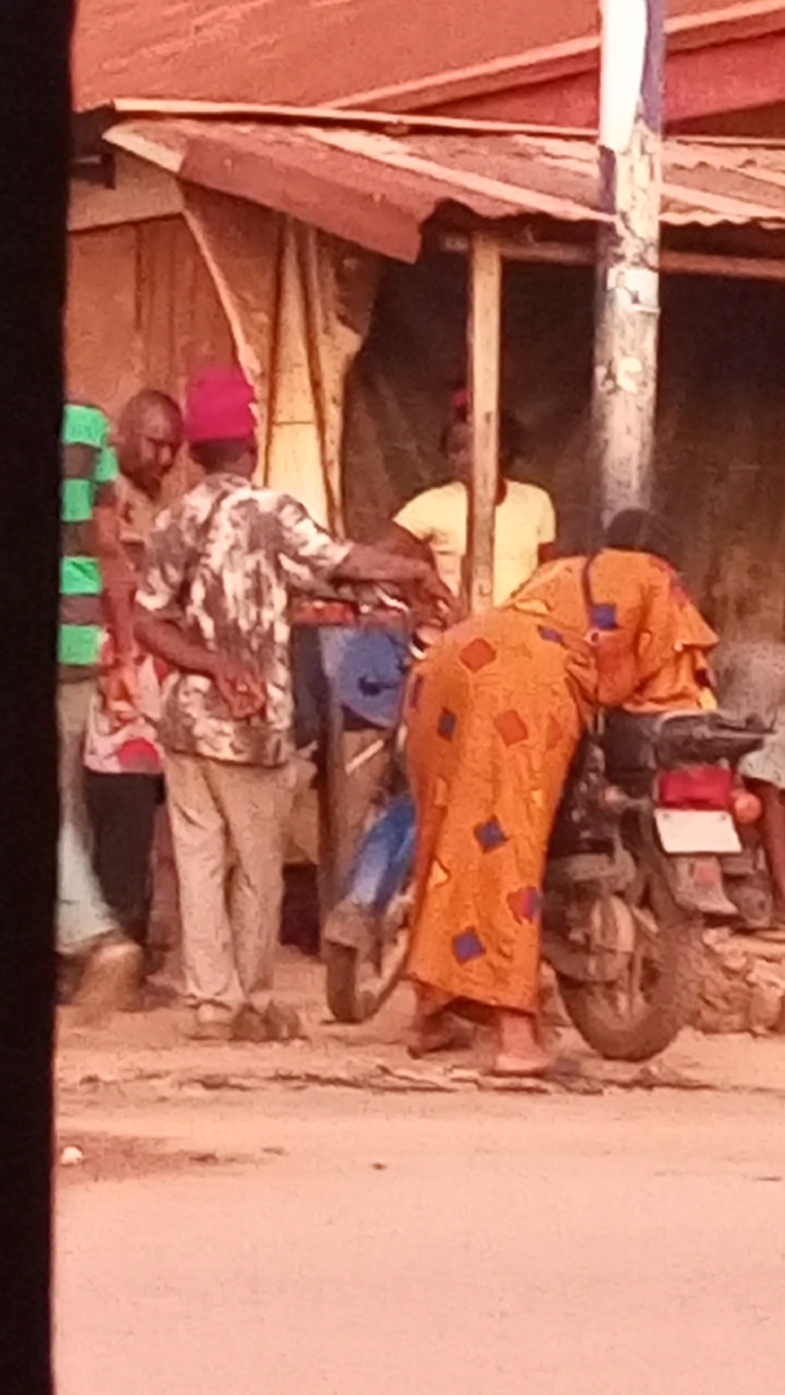 The image shows a group of people gathered around a motorcycle on a dirt road. One person, wearing a patterned orange garment, is bent over the motorcycle as if examining it. Several other people stand nearby. One man is wearing a patterned shirt and red hat. A building with a makeshift awning stands in the background. The location is indicated as Sabon Tasha, Nigeria.