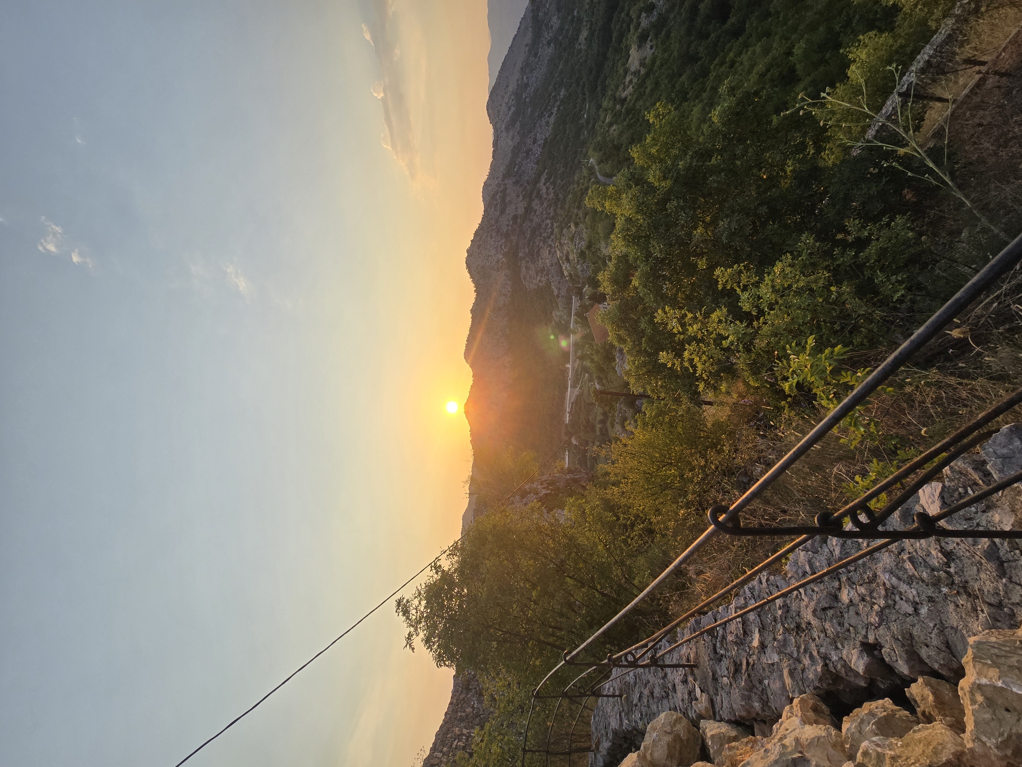 The image captures a sunset scene from an elevated vantage point in Gornja Vrbica, Montenegro. In the foreground, a stone pathway is bordered by a rustic metal railing. The railing is composed of dark metal poles with curved sections and some visible rust. The pathway is made of rough, uneven stones, with larger rocks present along the left edge.

Beyond the path and railing, a verdant hillside descends into a valley. Trees with lush green foliage dominate the midground, interspersed with rocky outcrops. A few scattered buildings with terracotta roofs are visible amidst the greenery. In the distance, a range of rugged mountains stretches across the horizon, their slopes covered in scrub and sparse vegetation.

The sun, a bright, glowing orb, is low in the sky, positioned just above a distant mountain peak. Its rays cast a warm, golden light across the landscape, creating long shadows and illuminating the sky with hues of orange, yellow, and pale blue. Wispy clouds are scattered across the upper portion of the sky. A power line stretches diagonally across the upper left quadrant of the image. The overall atmosphere is serene and natural, indicative of a rural mountainous environment during twilight.