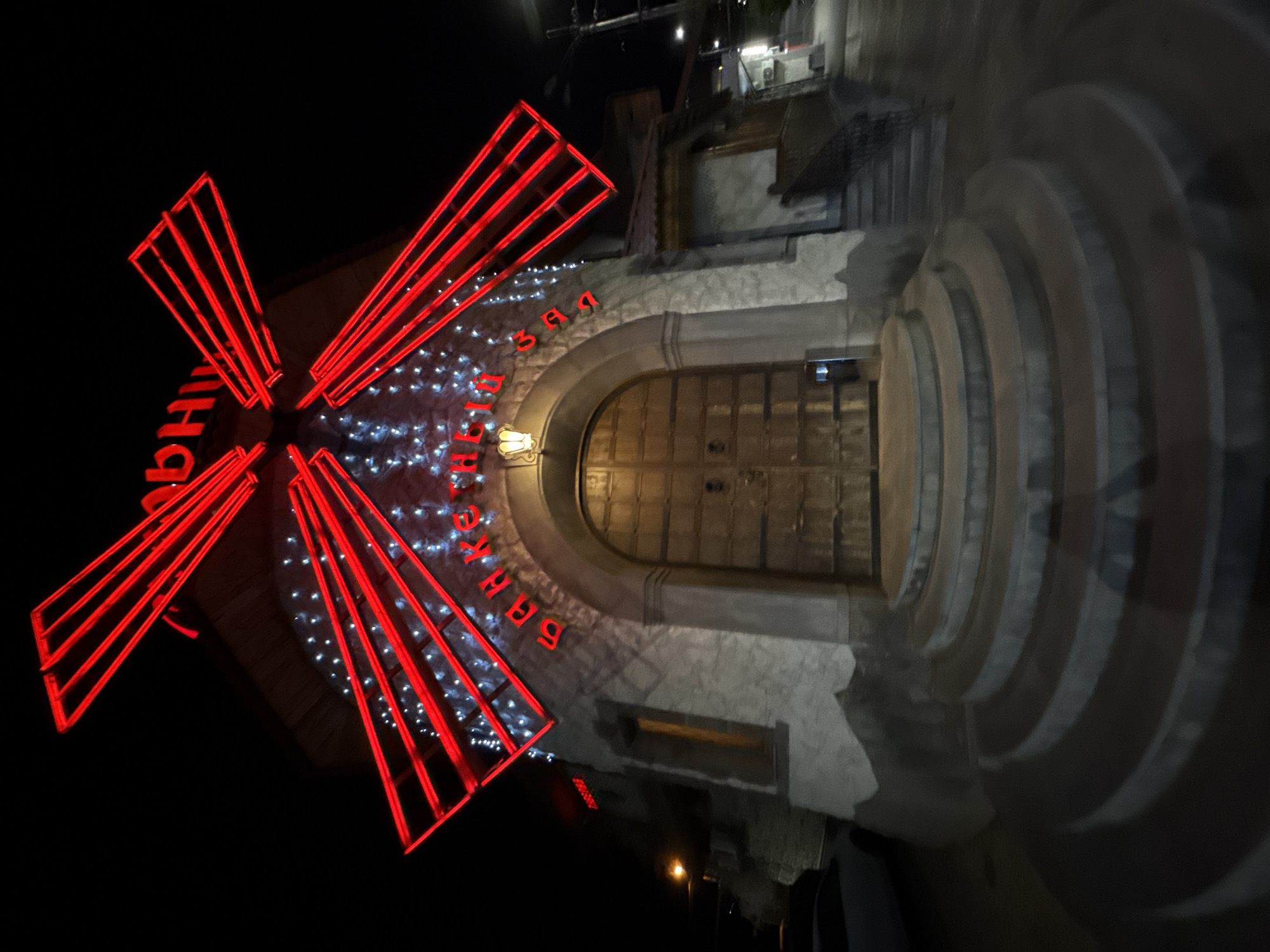 The image shows the "Moulin Rouge" entrance at night in an unknown city. The building features a prominent windmill structure on the left, lit with red neon. The entrance is defined by an arched doorway leading to a large wooden door. The building's facade is illuminated by white lights.
