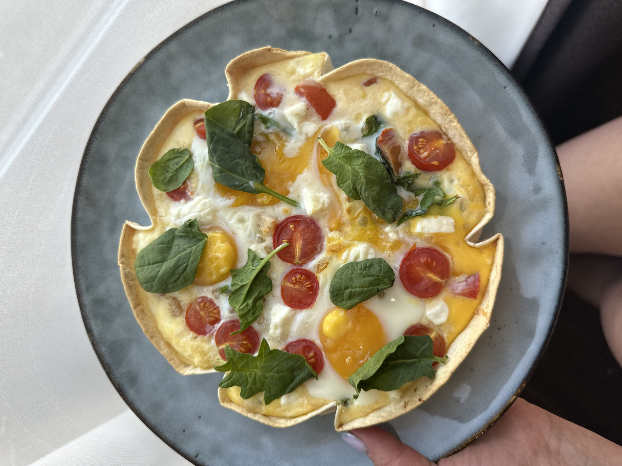 A close-up, eye-level shot shows a round, blue-grey plate with a slightly raised edge. On the plate is a baked tart with a light-colored, irregular crust. The tart filling consists of cooked eggs, sliced cherry tomatoes, small pieces of white cheese, and fresh spinach leaves. The tart is held by a person's hand, with part of their arm visible in the background. The background is blurred, showing a white surface and a dark object. The image's location is Chaikovsky, Russia.