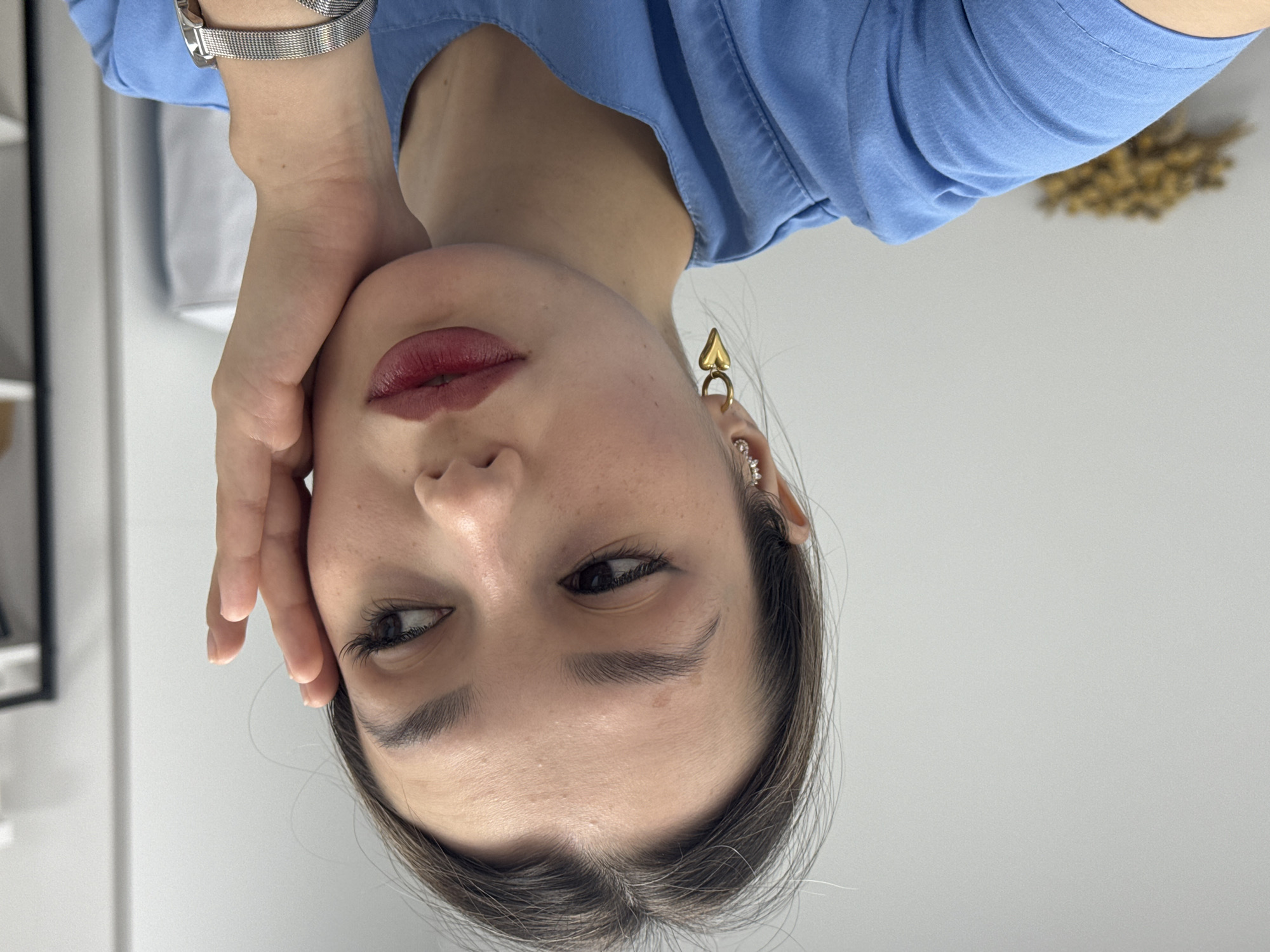 The image is a close-up portrait of a woman, rotated 90 degrees counter-clockwise so that she appears upside down. The woman has dark hair, fair skin, and a neutral, slightly pensive expression. She wears dark red lipstick, has well-defined eyebrows, and dark eyes with long eyelashes, looking towards the right side of the frame. Her left hand is gently placed against her left cheek and chin.
She is dressed in a light blue short-sleeved top. A silver mesh strap wristwatch is visible on her left wrist. On her right ear, she wears a distinctive gold ear cuff shaped like a spade, along with a smaller, sparkling earring higher up on her ear.
The background is a plain white or light grey wall, suggesting an indoor setting. In the upper right corner of the rotated image, a blurry golden-brown object, possibly dried flowers or wheat, is partially visible. A dark rectangular frame is also partially visible on the left side. There is no visible text in the image.