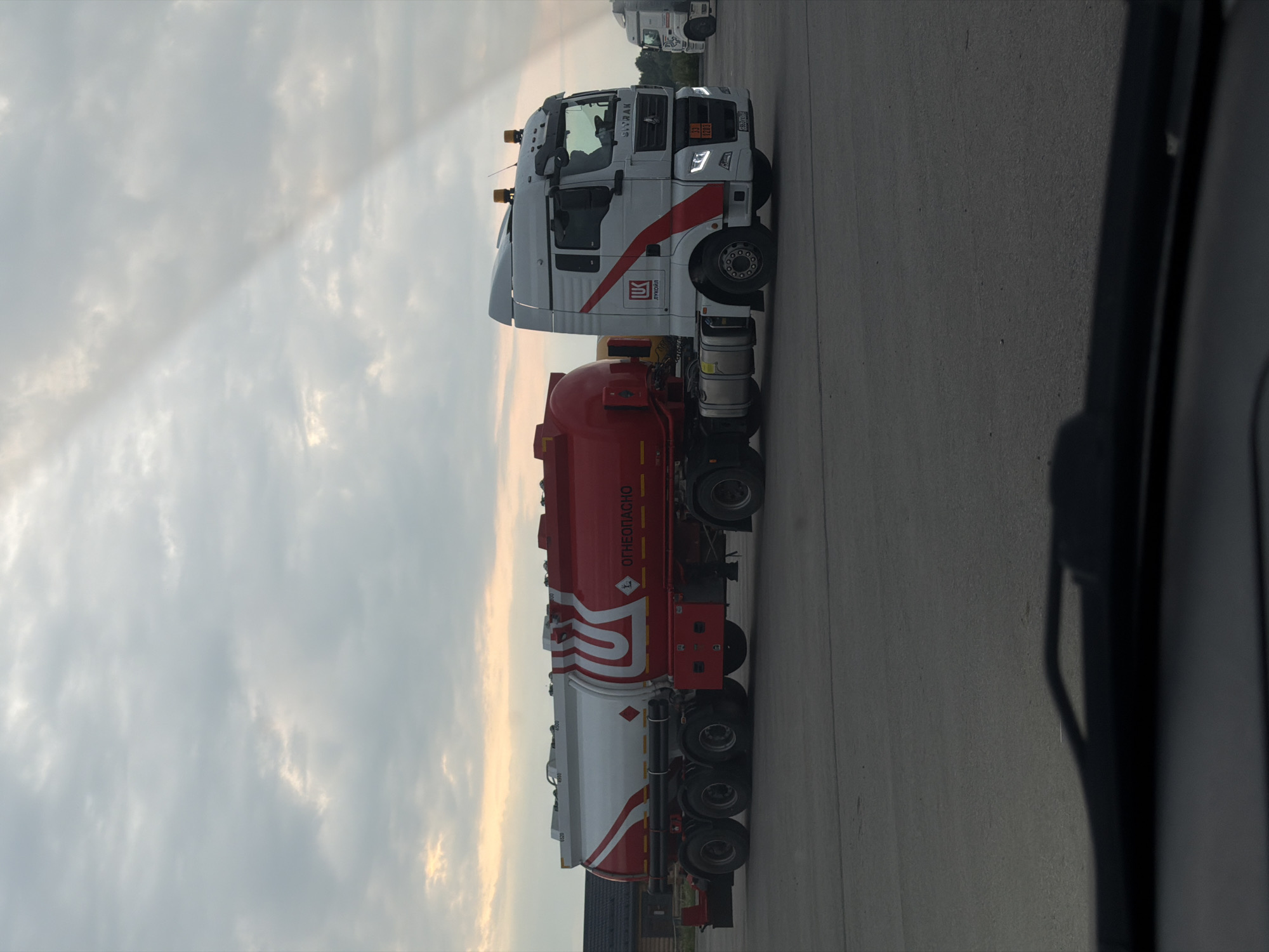A white and red tanker truck, branded with the Lukoil logo, is parked on a paved surface under a cloudy sky. The truck's trailer is predominantly red with a white section near the front, featuring the Lukoil emblem. Text on the side of the trailer reads "ОГНЕОПАСНО," which translates to "Flammable." The truck cab is white with red accents and has a license plate visible. In the background, a few other trucks are parked and a building with a dark roof is visible. The scene appears to be an outdoor lot, possibly a truck stop or depot, located in Усть-Сыны, Russia.