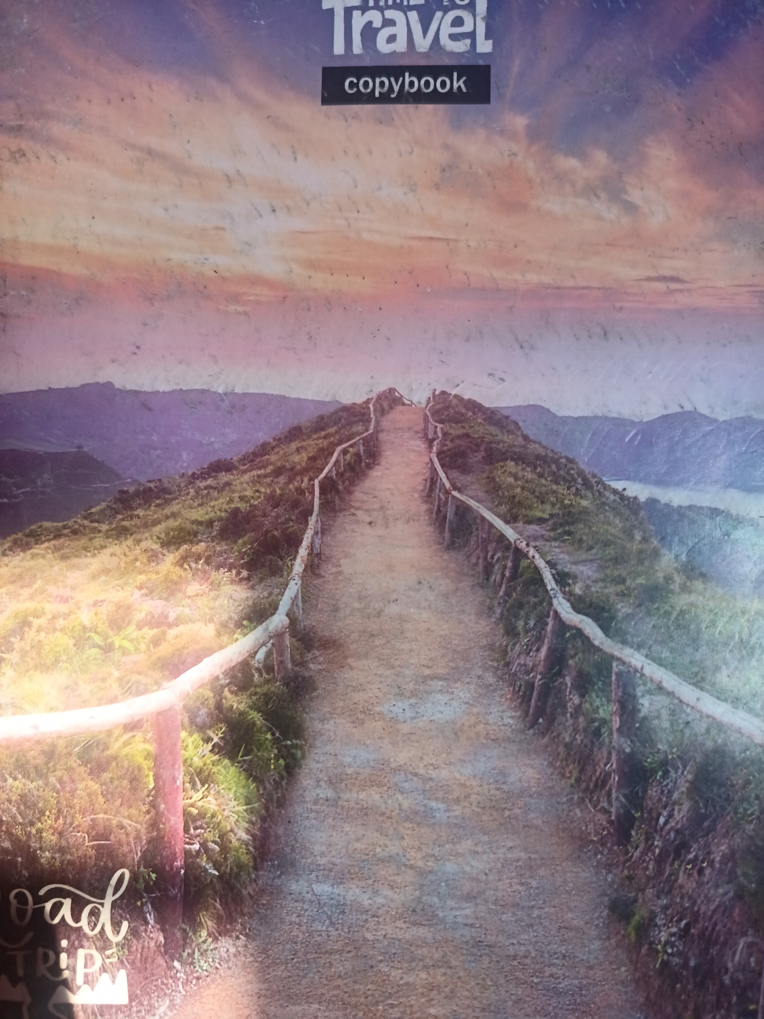 The image is a photograph of a dirt path winding along a ridge with wooden railings on either side. In the foreground, a sign with cursive lettering reads "Road Trip." The path leads up and away from the viewer, disappearing into the distance. The landscape is characterized by rolling hills covered in vegetation, with mountains in the background. The sky above is filled with clouds in shades of orange, pink, and purple, suggesting a sunset or sunrise. The overall impression is one of a scenic and adventurous journey. The image does not contain any information indicating it is located in Novokuznetsk, Russia.