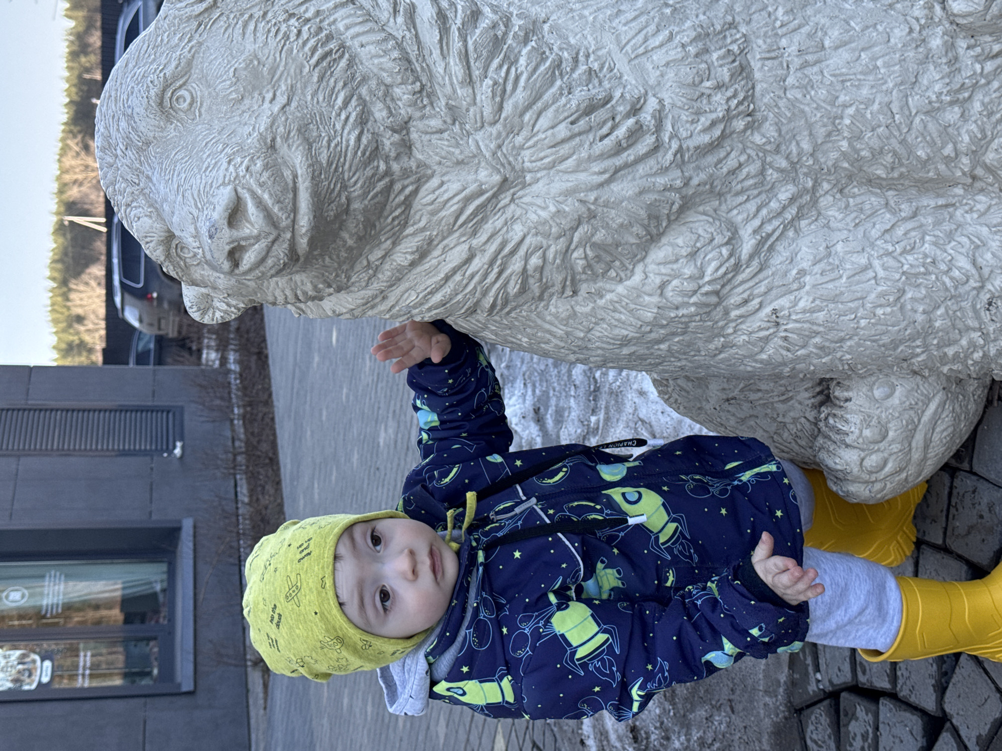 A toddler stands on a stone-paved surface next to a large, light-colored stone sculpture of a bear. The toddler wears a blue spacesuit-patterned jacket, gray pants, yellow boots, and a yellow hat with a matching pattern. The toddler is looking at the camera. The location is identified as Izhevsk, Russia. A building with glass windows is visible in the background. A vehicle is seen partially in the far background.