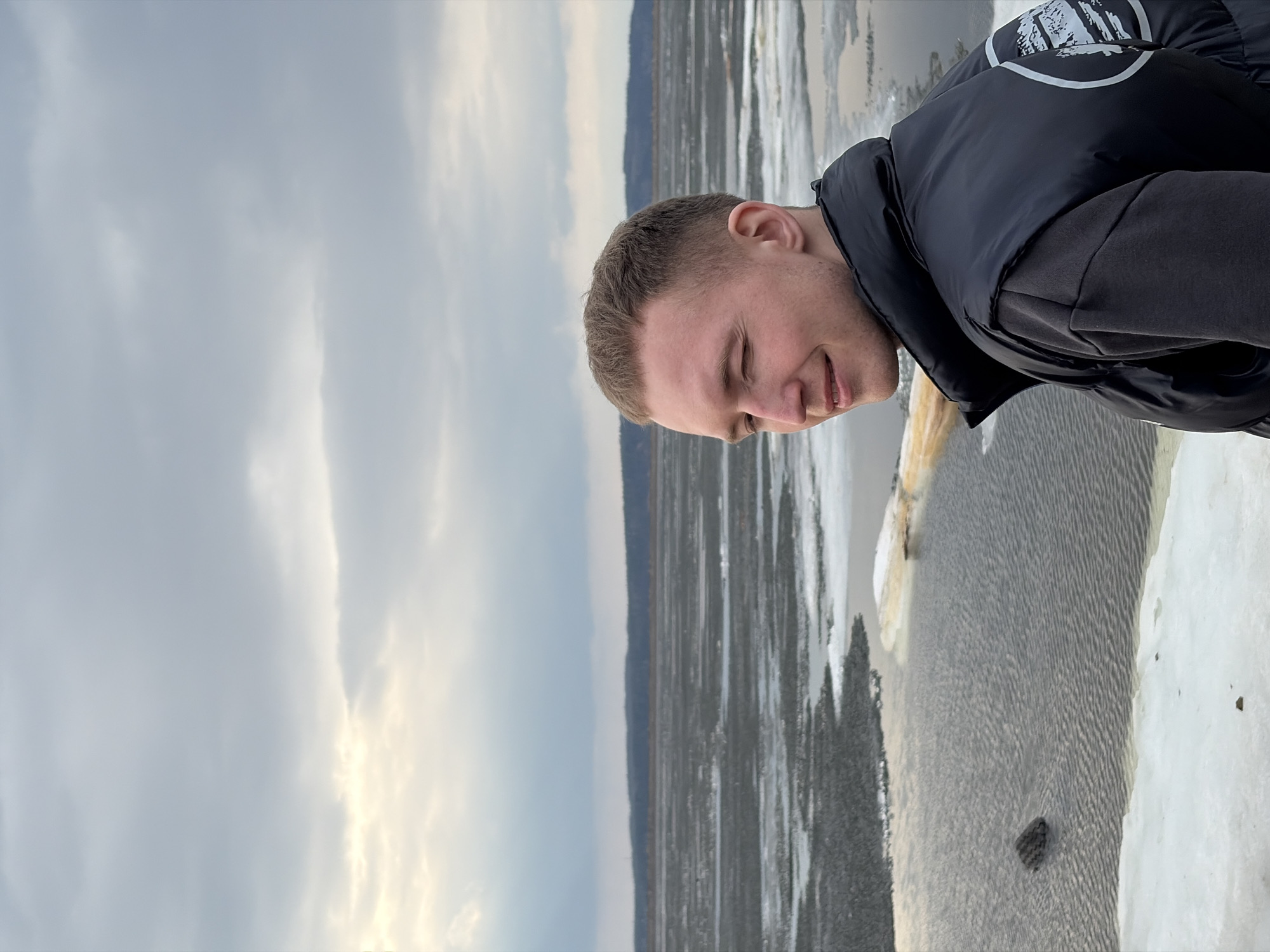 The image, taken in Chaikovsky, Russia, features a young man outdoors next to a partially frozen body of water, likely a river. The man, positioned slightly off-center, is shown from the chest up, with short, light brown hair and fair skin. He is wearing a black puffer vest over a dark long-sleeved shirt. He looks downwards and slightly to his right, with his eyes mostly closed and a subtle smile or smirk on his face, suggesting amusement or contemplation.
To the man's right, the body of water exhibits ripples and scattered ice fragments, indicating a cold environment. The bank on his immediate left is lined with snow and ice. In the distance, across the water, a dark, low-lying landmass, possibly a forest, is visible beneath an overcast sky. The sky is predominantly gray with some lighter, diffused patches of sunlight, suggesting a cloudy daytime. The cold weather is further reinforced by the man's warm clothing and the icy conditions. A white, stylized graphic or logo is visible on the back of his vest, though its specific details are not clearly legible.