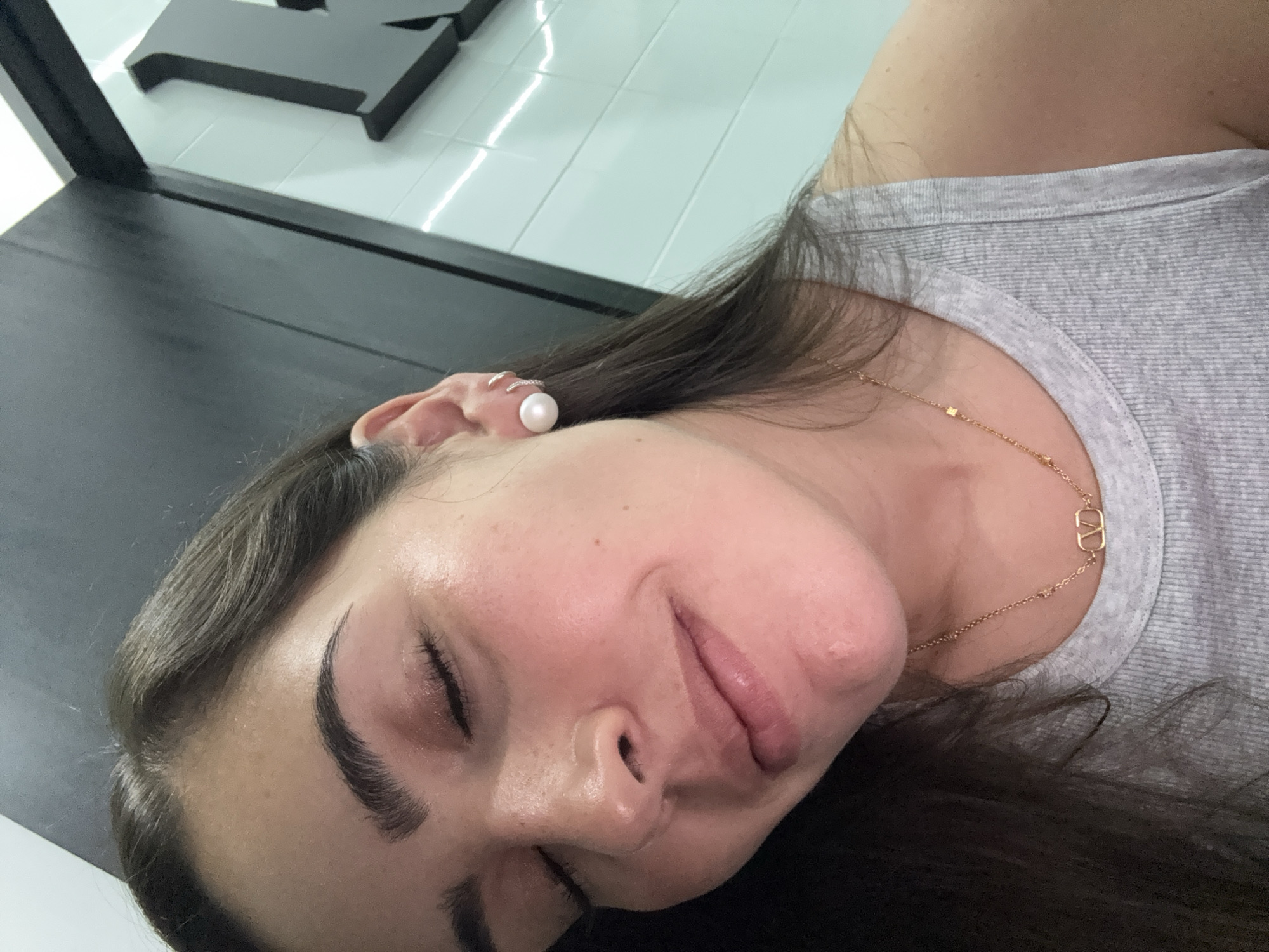 A woman lies with her eyes closed. She has dark hair and light skin. She wears a pearl earring and a gold necklace. She appears to be lying on a black surface. The background is out of focus and shows white tiles and a black object. The location is unknown.