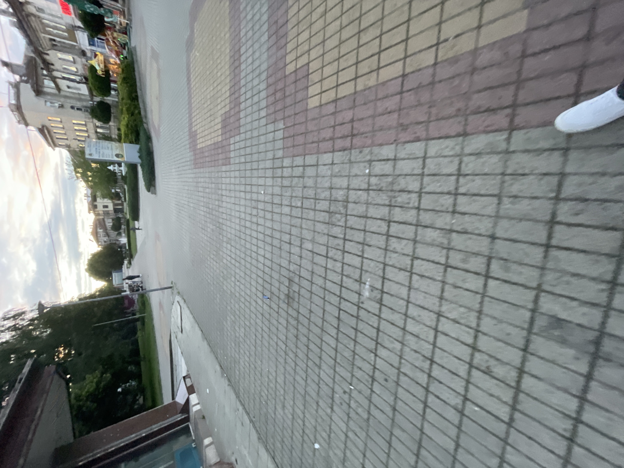 The image presents a ground-level view of a paved street or town square in Ihtiman, Bulgaria, observed during the late afternoon or early evening. The sky, visible towards the upper left, is partly cloudy with a warm, soft light near the horizon.
In the foreground, a wide sidewalk dominates, constructed from square pavers arranged in a geometric pattern of grey, pinkish-brown, and yellowish-beige sections. Small pieces of litter, including white paper scraps and a tiny blue object, are scattered on the grey pavement. A white shoe is partially visible on the far right side, indicating the presence of the photographer.
Beyond the sidewalk lies an open urban space, possibly a street or pedestrian area. On the left, there is a strip of greenery with bushes and trees. A tall, thin grey pole stands in the midground. In the distance, blurry figures of people and what might be a vehicle suggest typical street activity.
The background features several multi-story buildings characteristic of European architecture. One building on the left exhibits traditional facades with visible windows that reflect a warm, golden light. Above another building, a red sign with partially visible white text, likely reading "...TEL", indicates a commercial establishment, possibly a hotel. Closer to the buildings and trees, a rectangular white sign mounted on a pole displays text in Cyrillic script, serving as an informational board for the area. The scene conveys a quiet, everyday urban environment.