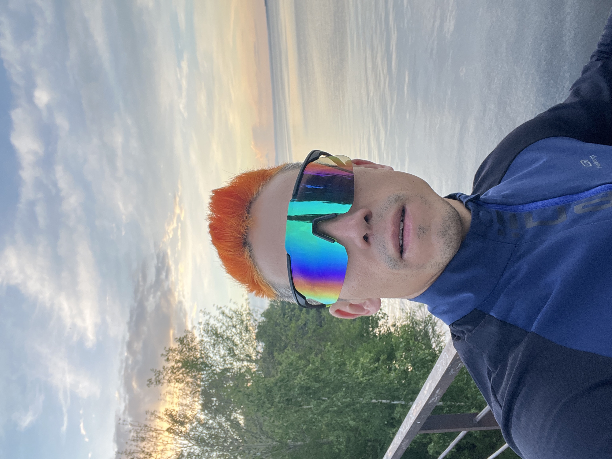 A selfie-style, tilted, full-frame photograph depicts a man with bright orange hair wearing iridescent sunglasses. The man is wearing a dark blue and black long-sleeved jacket with a visible "Kalendji" logo on the chest. He appears to be outdoors, standing on a metal railing or bridge, with trees and a body of water visible in the background. The sky is partly cloudy with warm sunset hues. The exact location in the city of Chaikovsky, Russia, cannot be determined with certainty from the visual information provided.