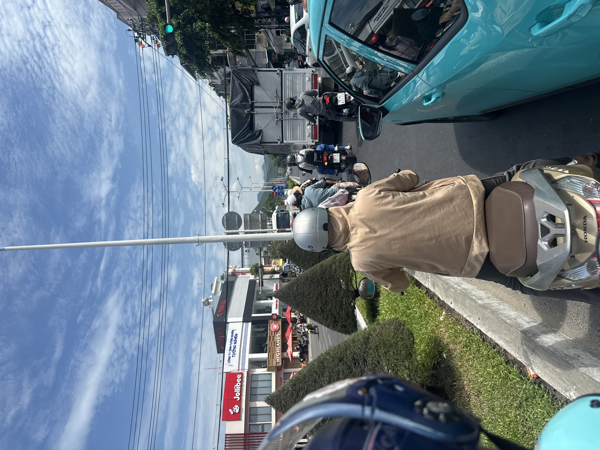 The image captures a street scene in Nha Trang, Vietnam, with a procession of motorbikes and a truck navigating a roadway under a partly cloudy sky. The perspective is from the back of a motorbike, with a rider wearing a light brown hooded jacket and a silver helmet visible in the foreground. To the right, the teal-colored car, possibly a Hyundai Accent, is partially visible. Across the street, a Jollibee restaurant and a Yamaha showroom are identifiable landmarks, with manicured conical bushes lining the median. The presence of multiple motorbikes, a large truck, and the architecture of the buildings suggests a bustling urban environment. Traffic signals and power lines overhead further indicate an active street. The scene appears to be during daylight hours, with bright sunlight casting shadows.
