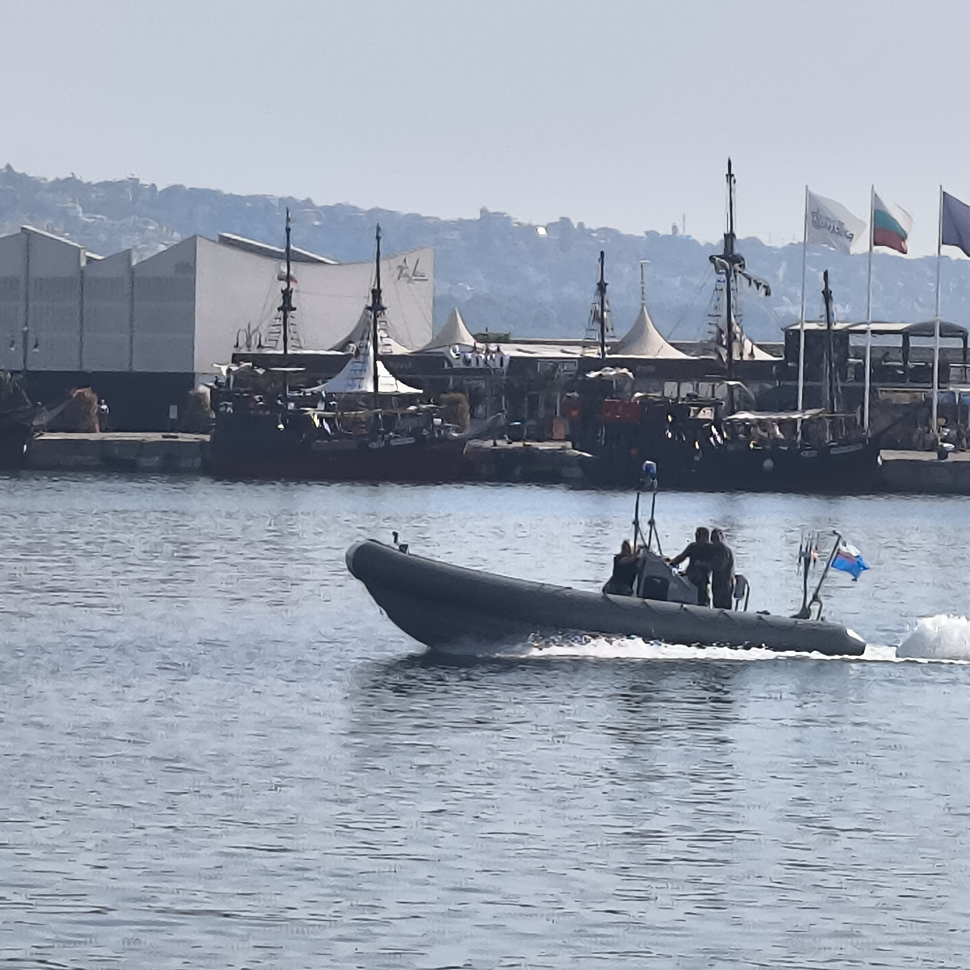 In Varna, Bulgaria, a dark grey inflatable boat with two individuals on board travels at speed across calm blue water. The boat is equipped with a flag at its stern, and a blue light is visible at the front of the vessel. In the background, large, dark ships with masts and white sails are docked along the shoreline, adjacent to industrial-looking buildings and tents. The shoreline itself is lined with structures and backed by a light-colored, rocky hillside. The sky is a pale, hazy blue.